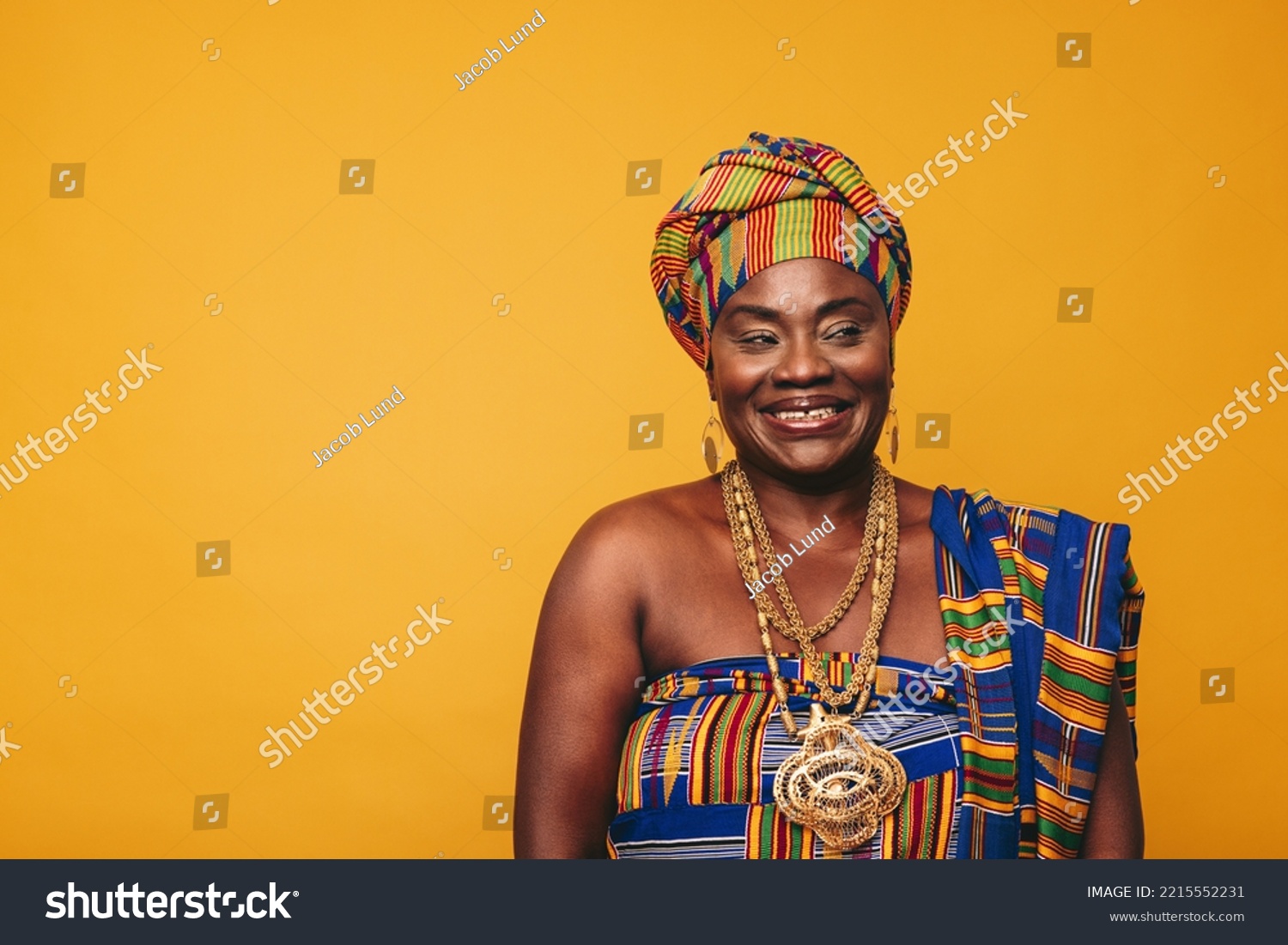 Smiling Ghanaian woman wearing a traditional attire against a yellow background. Happy black woman dressed in colourful Kente cloth and golden jewellery. Mature elegant woman embracing her rich cultur #2215552231