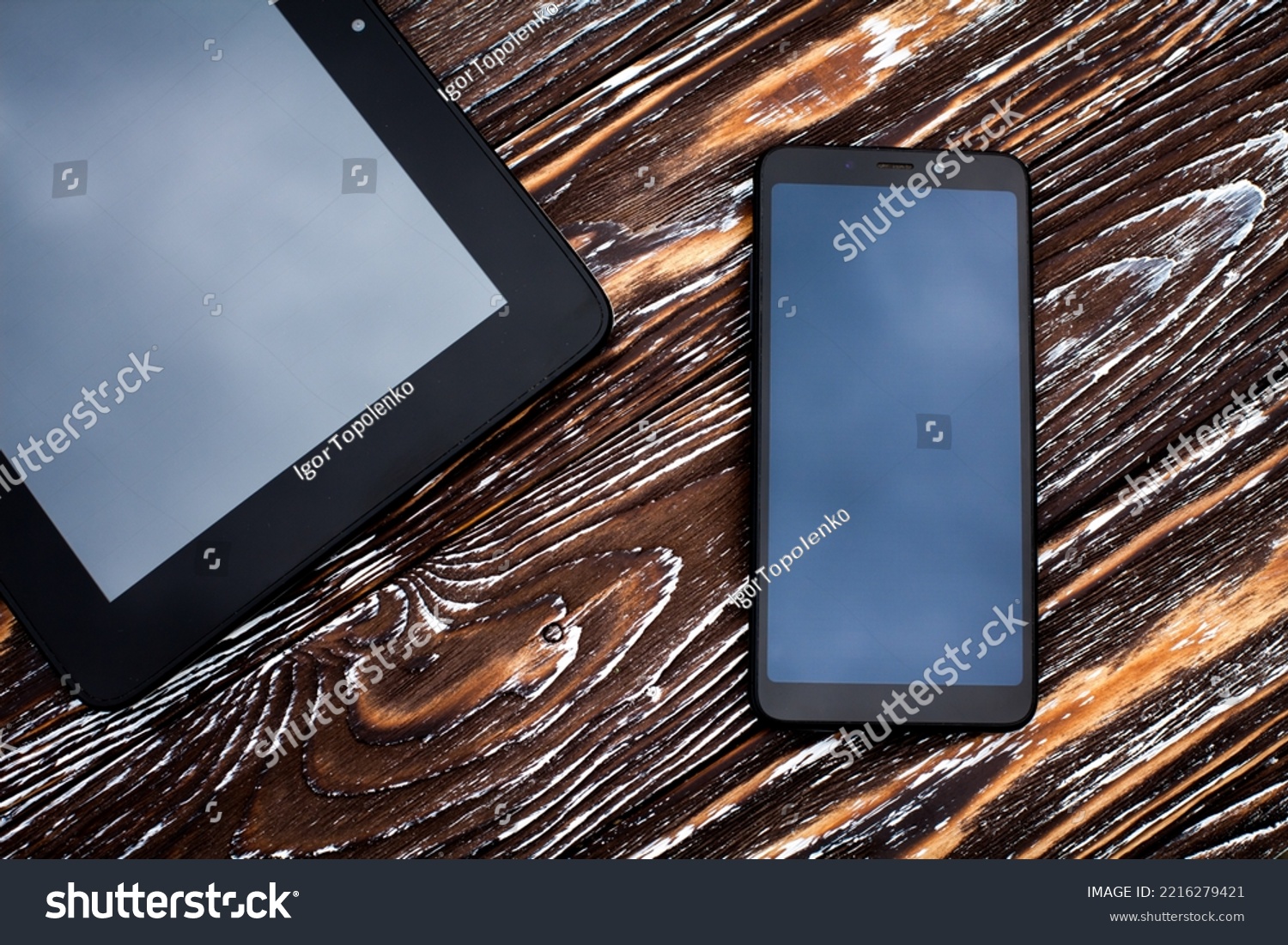 Two black devices, a tablet and a mobile phone lie on a dark wooden textured surface. #2216279421