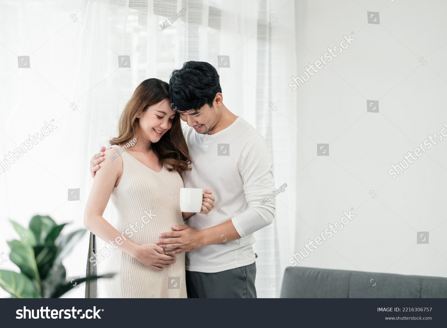 A happy asian couple. A beautiful pregnant wife is holding a cup of milk for the baby with her husband smiling and both of their hands rubbing the wife's tummy in the living room. #2216306757