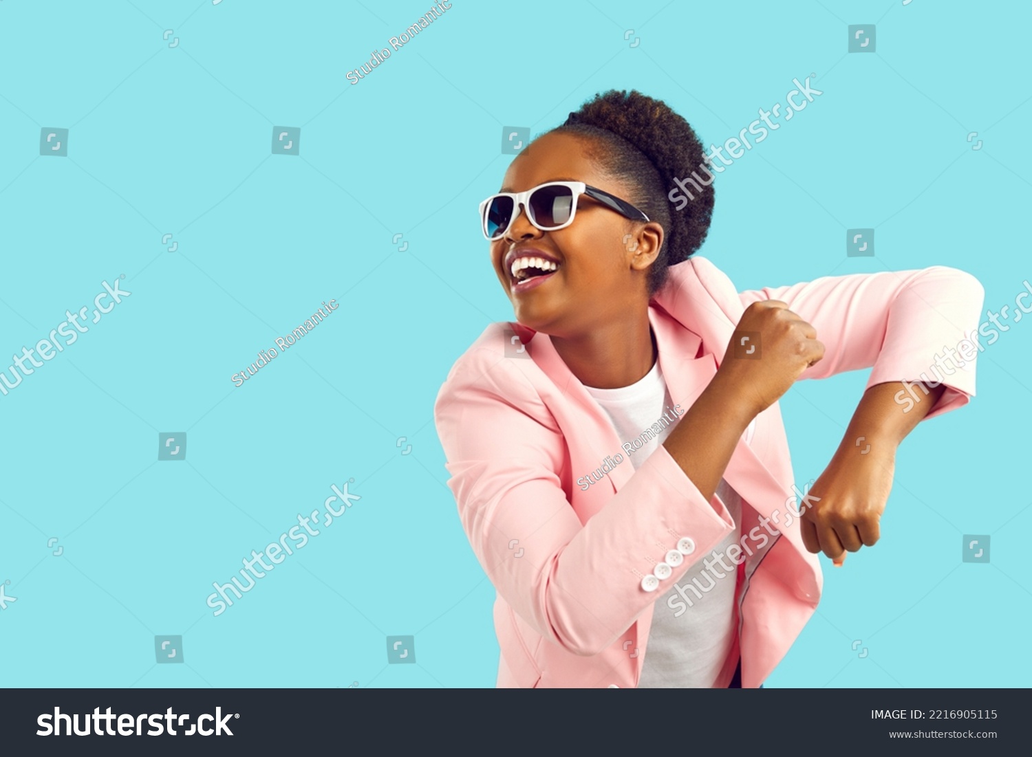 Cheerful happy excited young African American girl in pink suit and sunglasses dancing and having fun. Portrait of funny woman dressed up for party dancing isolated on bright blue copyspace background #2216905115