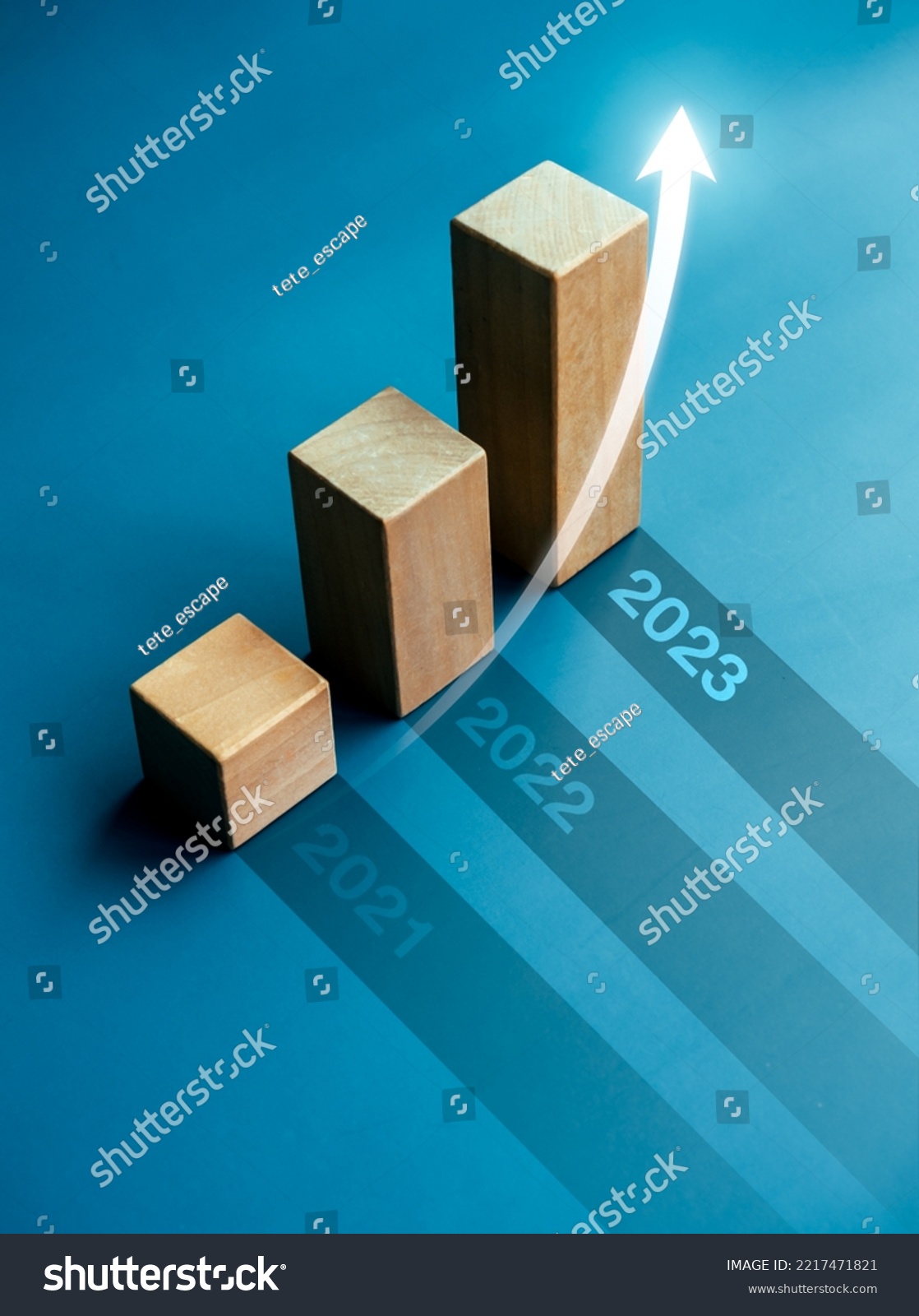 Shining rise up arrow on wooden cube blocks, bar graph chart steps on blue background with year numbers, 2023, 2022, 2021, vertical style. Business growth and development to success concepts. #2217471821