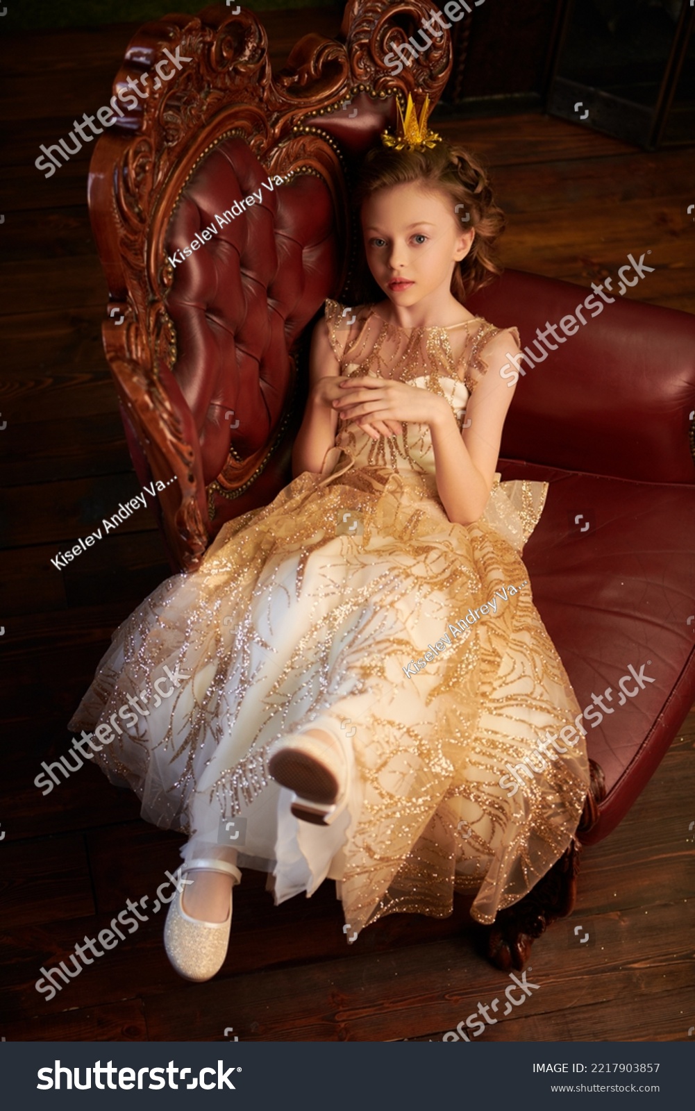 Fairy tales. Beautiful little princess girl in a golden dress and a crown sits in an armchair in her vintage castle. Full length shot.  #2217903857