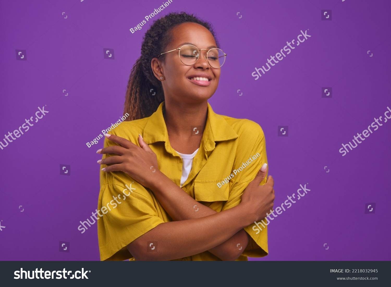 Young happy beautiful African American woman millennial with curly hair hugs herself to cheer up and get rid of loneliness dressed in casual yellow shirt, stands on isolated purple background. #2218032945