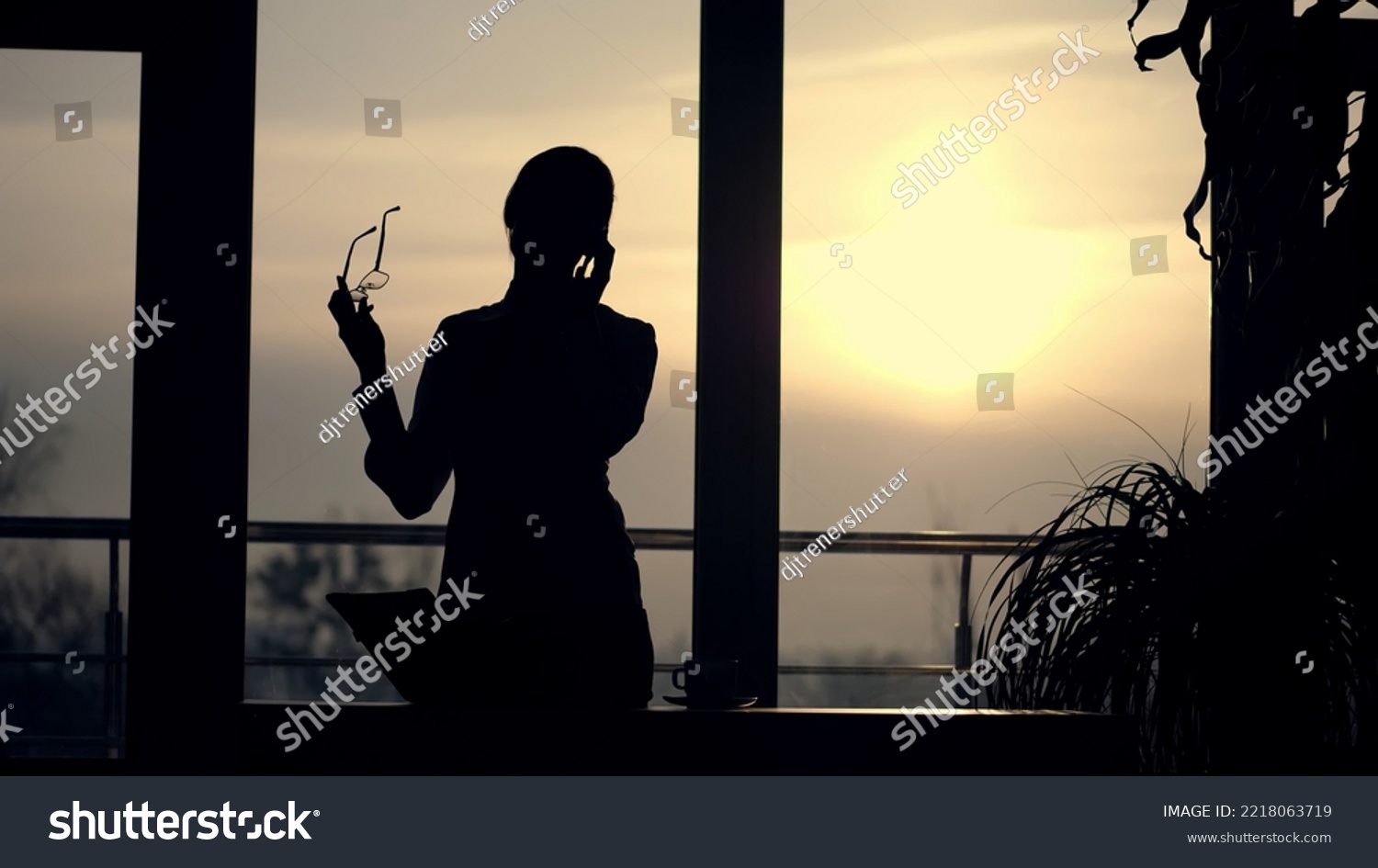 dark silhouette of Young business woman. she holds glasses in her hand, talks emotionally on mobile phone, gesticulates with hands, against background of large office window, at sunset, in rays of #2218063719