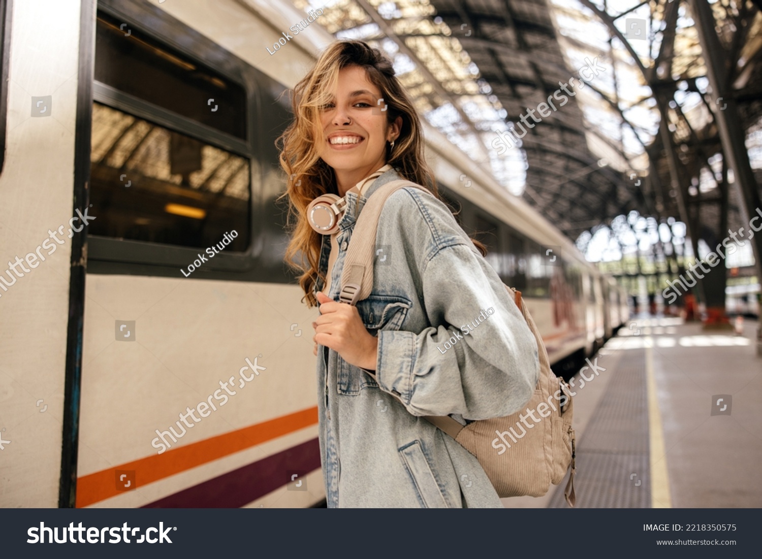 Happy young caucasian woman smiles with teeth looking at camera going on train trip. Blonde wears denim jacket, backpack. Spring break concept. #2218350575
