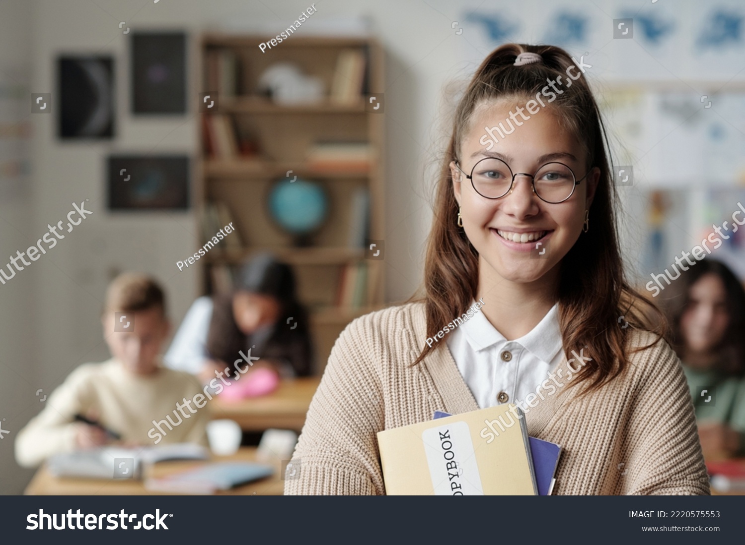 Smiling youthful schoolgirl in eyeglasses and casualawear looking at camera while standing in front of her classmates at lesson #2220575553