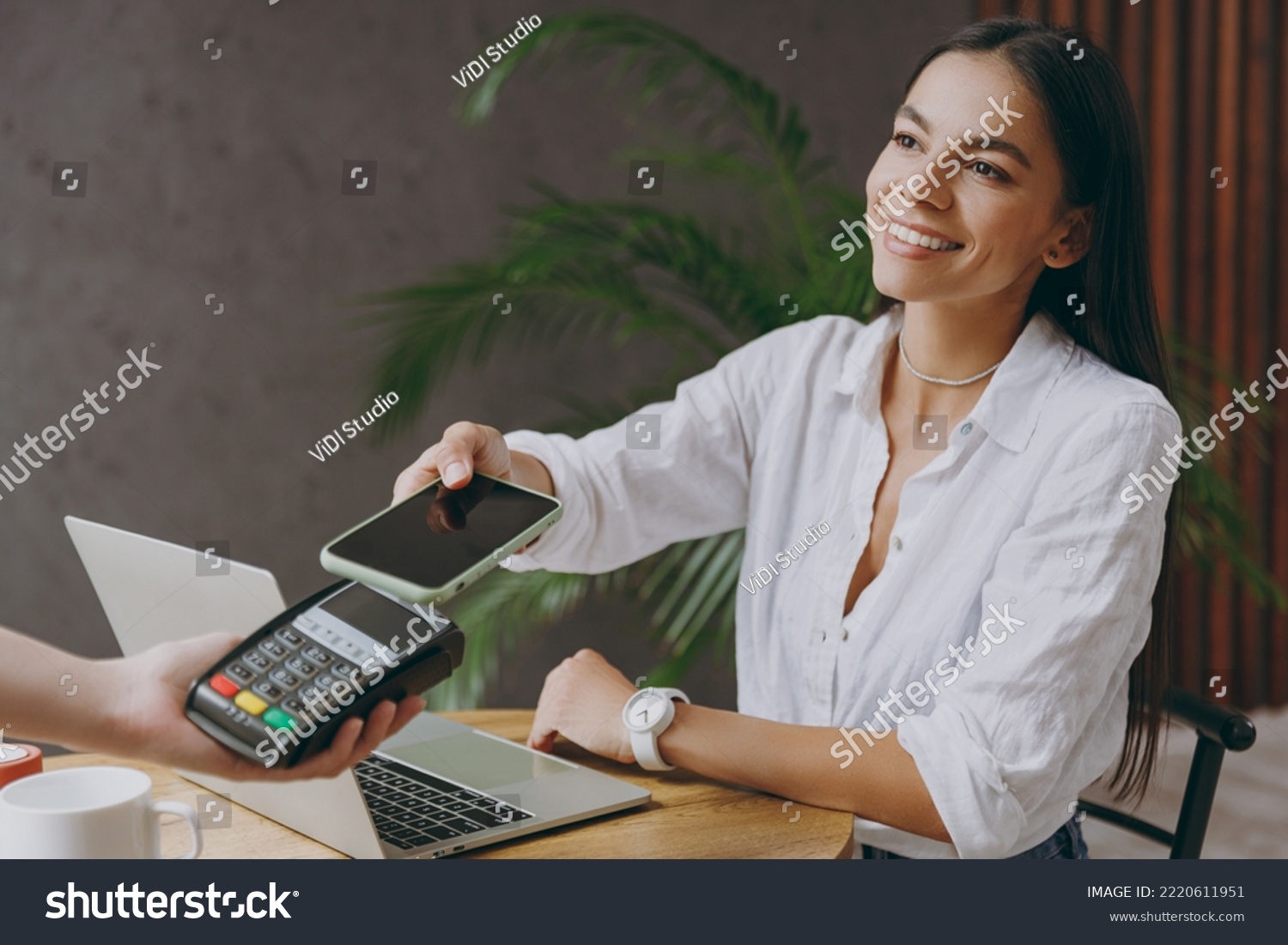 Young happy woman wear white shirt hold pay waiter hold mobile cell phone wireless bank payment terminal waiter sit at table in coffee shop cafe restaurant indoor work or study on laptop pc computer #2220611951