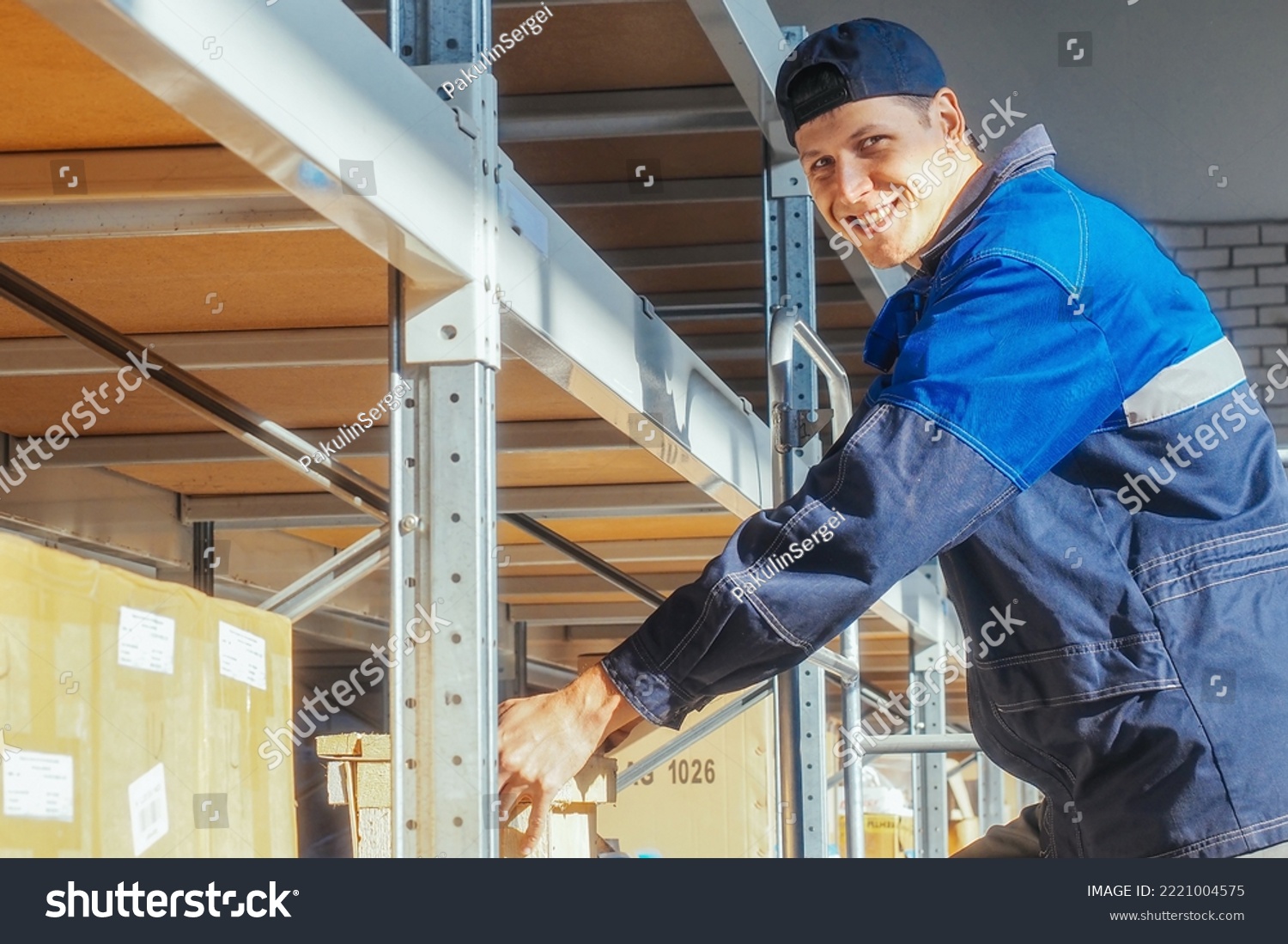 Storekeeper puts box on rack shelf looks at camera and smiles. Young Caucasian warehouse worker in overalls and baseball cap lays out goods inside warehouse. Authentic workflow. #2221004575