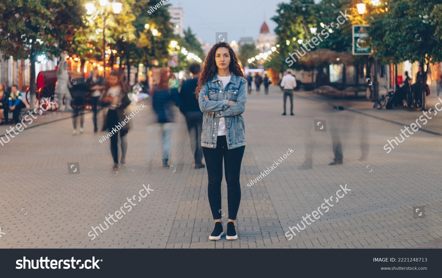 Portrait of stylish young lady tired of usual haste standing in the street among whizzing people and looking at camera. Time, youth and modern society concept. #2221248713