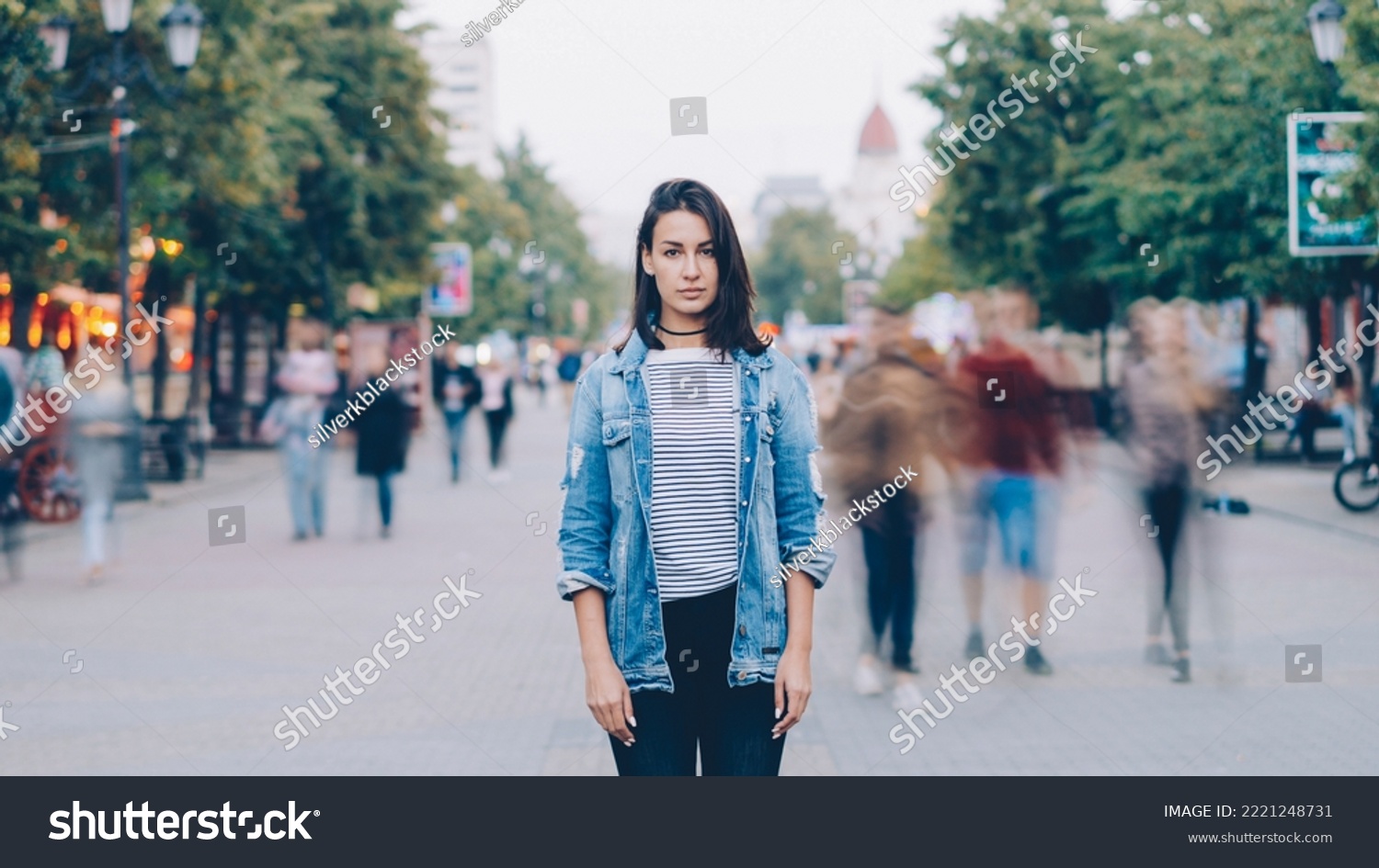 portrait of tired young woman student standing alone in city center and looking at camera with straight face while crowds of men and women are whizzing around. #2221248731