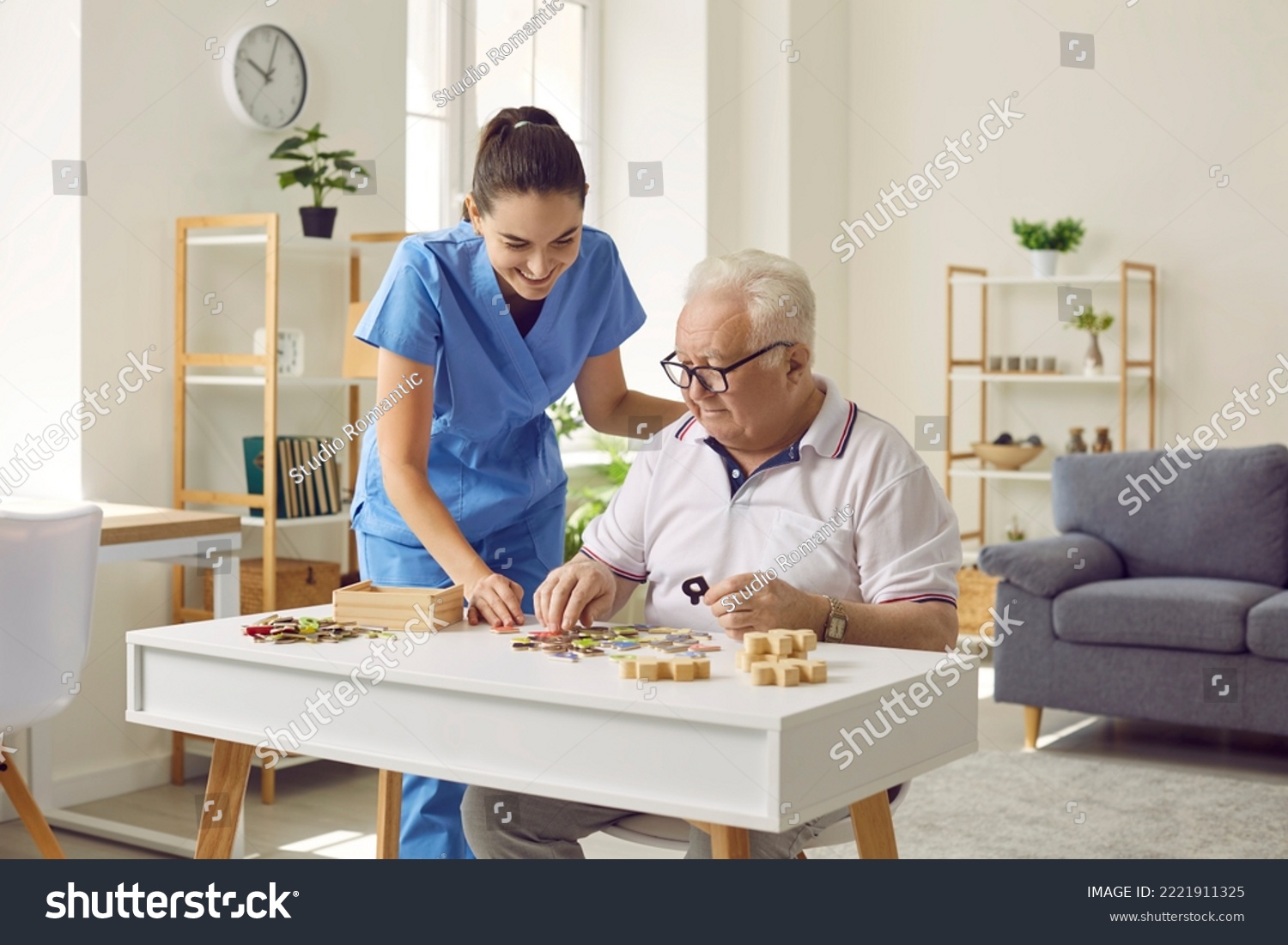 Smiling young female social worker and old man playing with jigsaw puzzle in nursing home. Caregiver in medical uniform helps elderly man to put together puzzles and letters. Old age concept. #2221911325