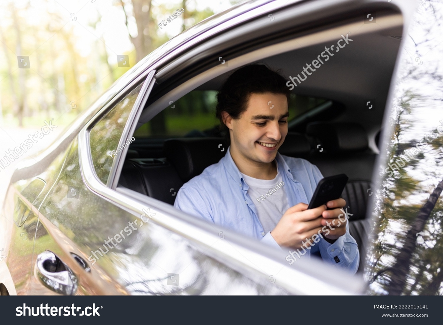 Happy smiling business man typing message on phone while sitting in a taxi. Young businessman in formal clothing using smartphone while sitting on back seat in car. Cheerful guy messaging with cell. #2222015141