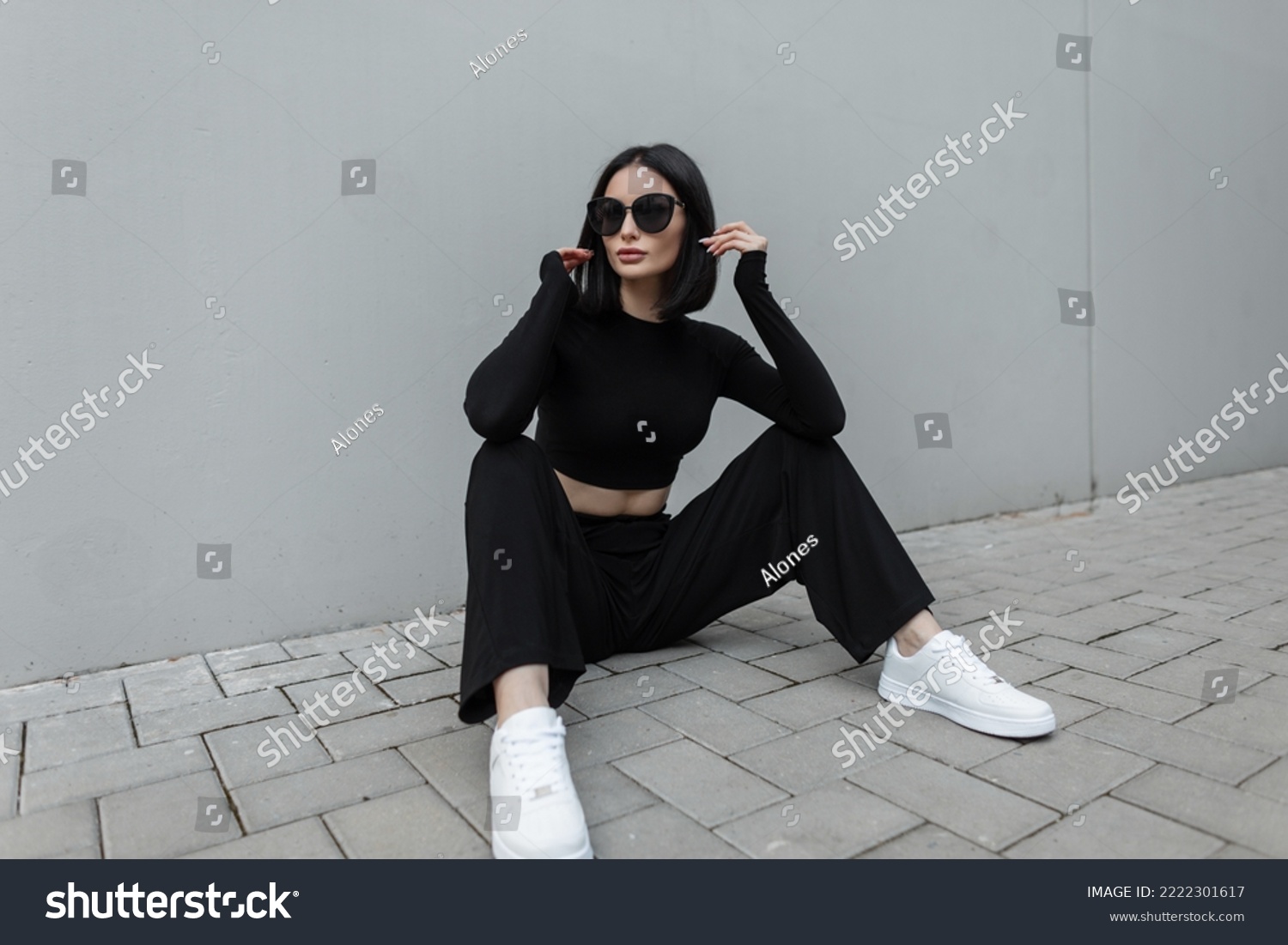 Fashionable young urban woman with black trendy sunglasses in fashion black stylish sports outfit with white sneakers sits on the street near a modern gray building #2222301617