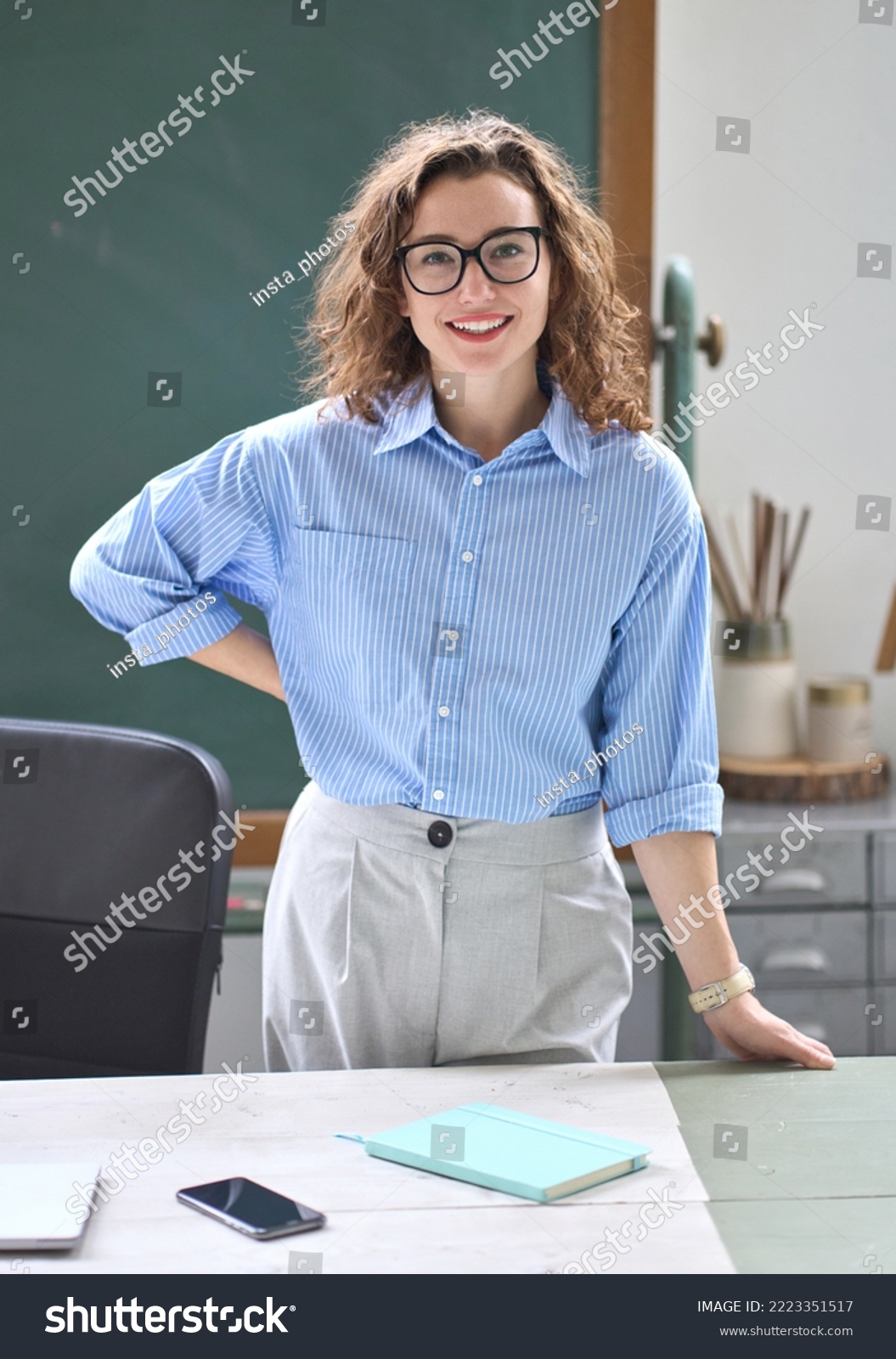 Young happy woman school professional teacher, female instructor coach standing at desk in front of chalkboard in classroom working in office presenting business education training, vertical portrait. #2223351517