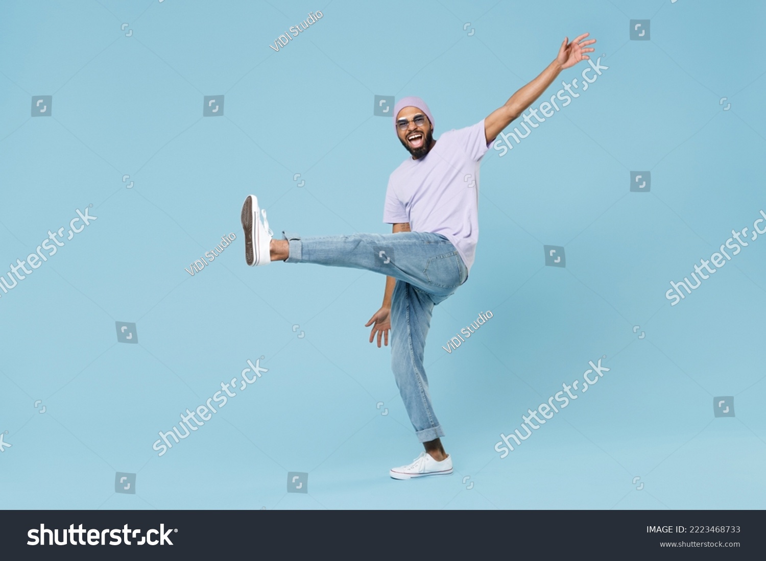 Full body young smiling happy man of African American ethnicity 20s wearing violet t-shirt hat glasses standing with raised up leg outstretched hands isolated on pastel blue background studio portrait #2223468733
