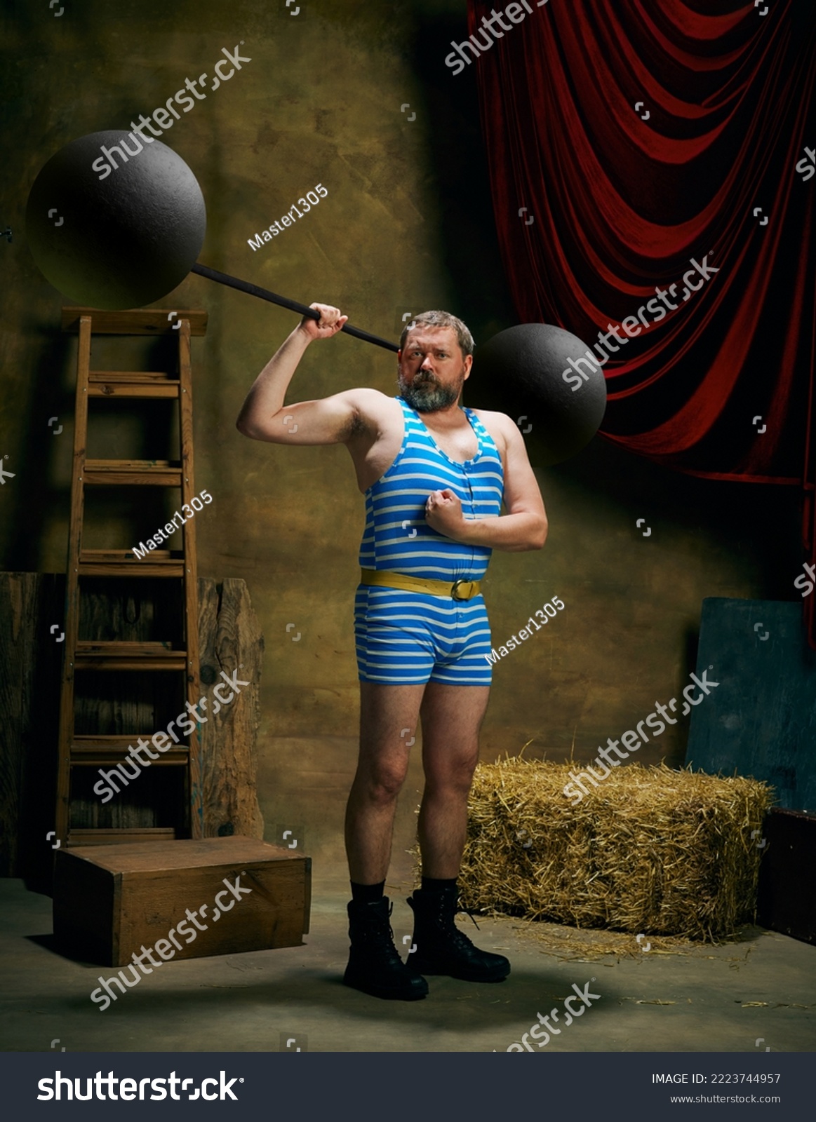 Retro circus. Cinematic portrait of retro circus strongman wearing striped sports swimsuit holding barbell on dark circus backstage background. Concept of creative art, fashion, style and inspiration #2223744957