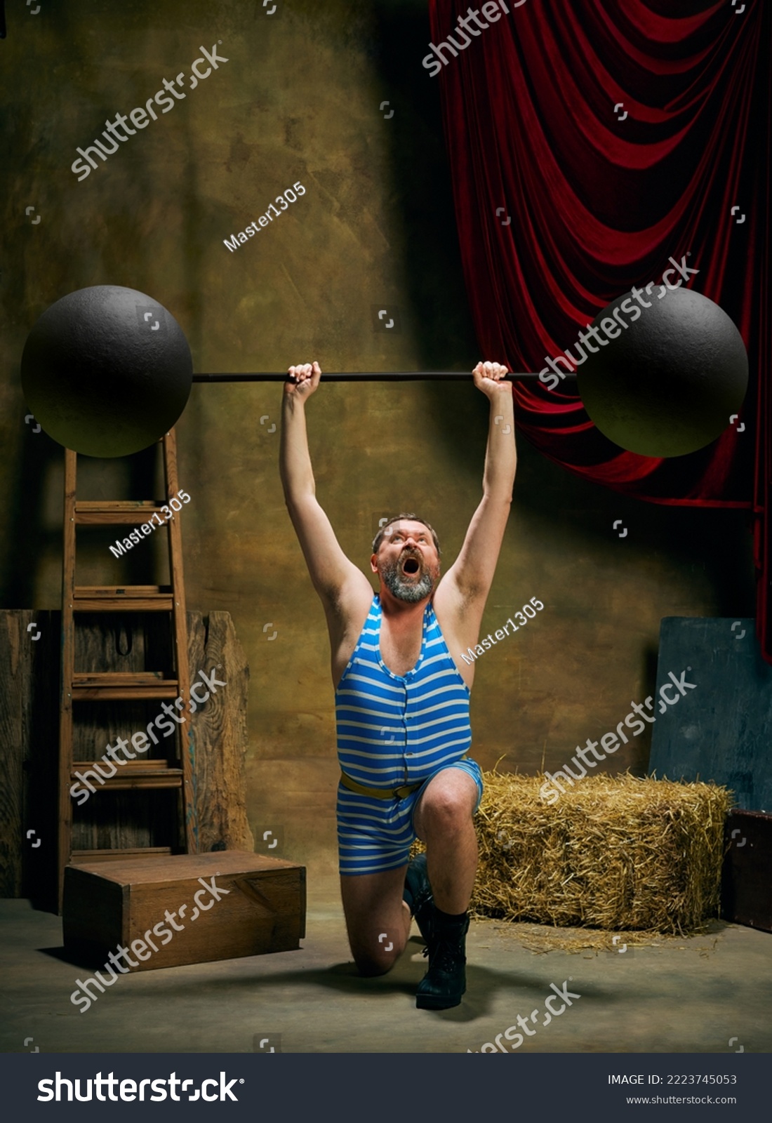 Circus athlete. Vintage portrait of retro circus strongman wearing blue striped sports swimsuit training with barbell over dark circus backstage background. Concept of creative art, fashion, style #2223745053