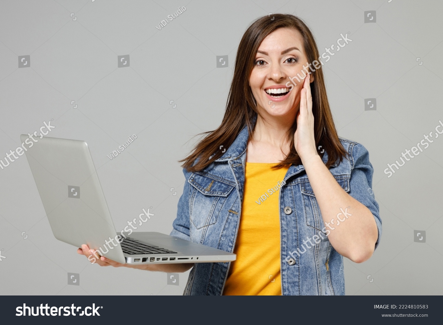 Young fun surprised caucasian woman 20s wearing casual trendy denim jacket yellow t-shirt point index finger on laptop pc computer browsing hold face isolated on grey color background studio portrait #2224810583