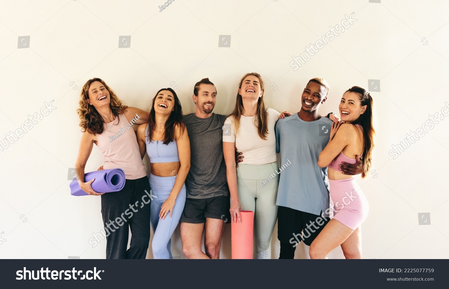 Group of happy fitness friends laughing while standing against a wall with yoga mats. Multicultural people attending a yoga class together. Sporty people working out in a community fitness studio. #2225077759