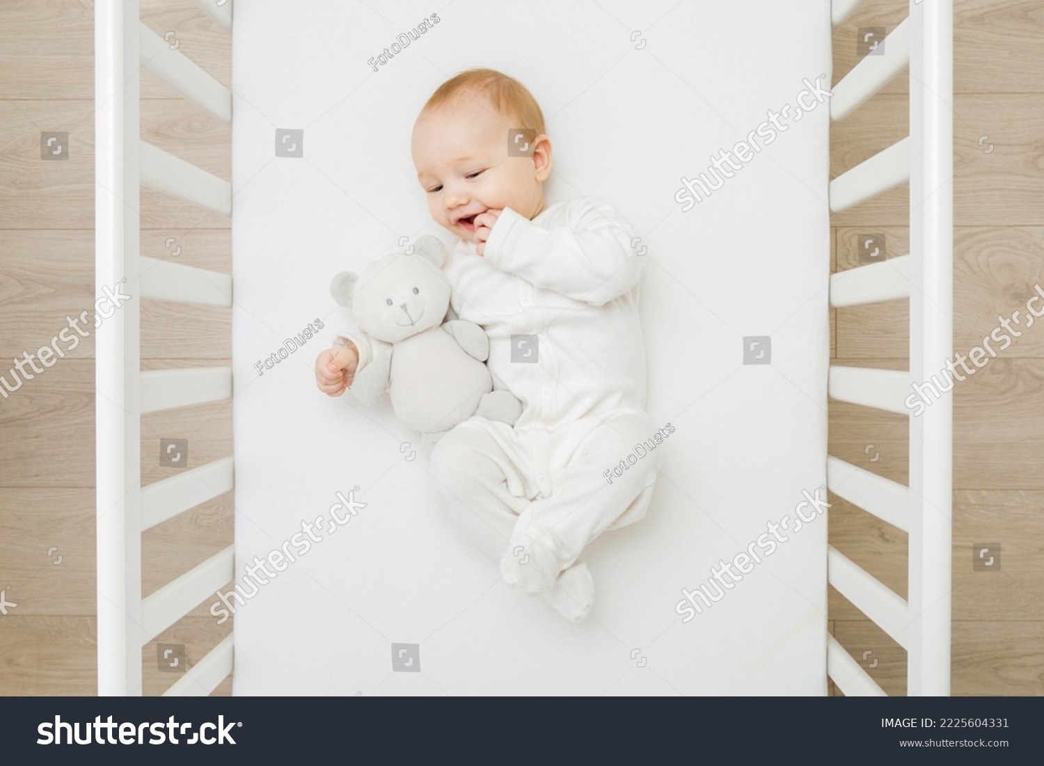 Adorable happy baby boy in white bodysuit hugging teddy bear and  lying down on back on mattress in crib at home room. Top view. 6 months old infant playing with first friend. Closeup.  #2225604331