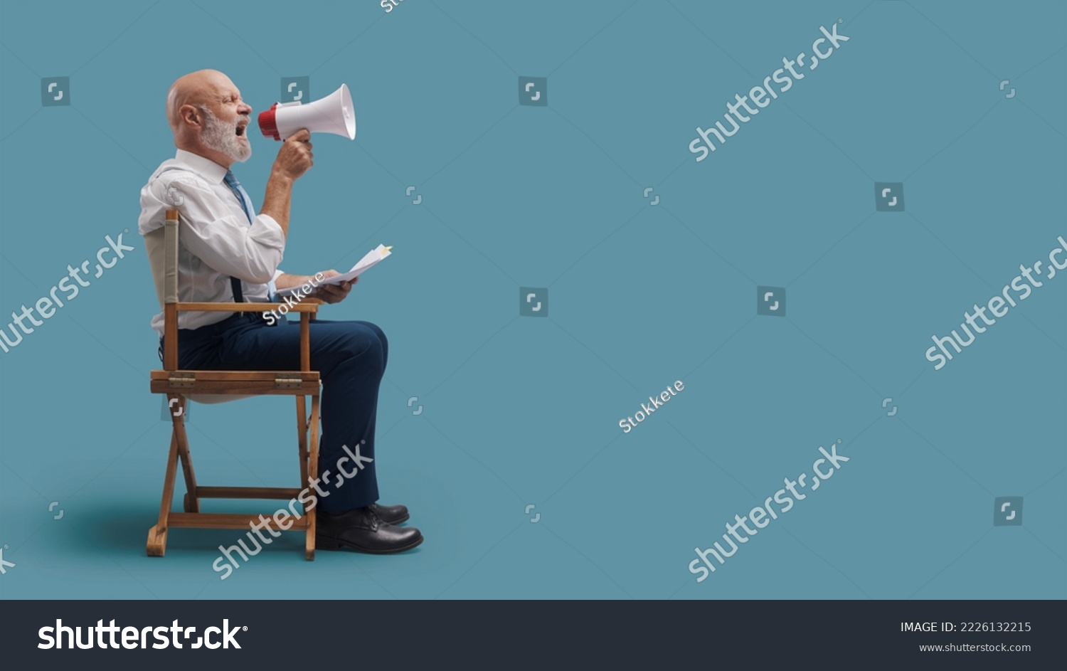 Confident professional film director sitting on the director's chair and shouting with a megaphone, film industry concept #2226132215