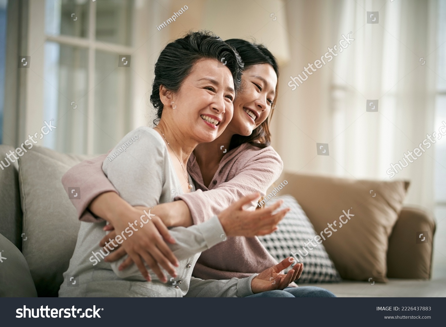 loving asian adult daughter sitting on couch at home hugging senior mother happy and smiling #2226437883