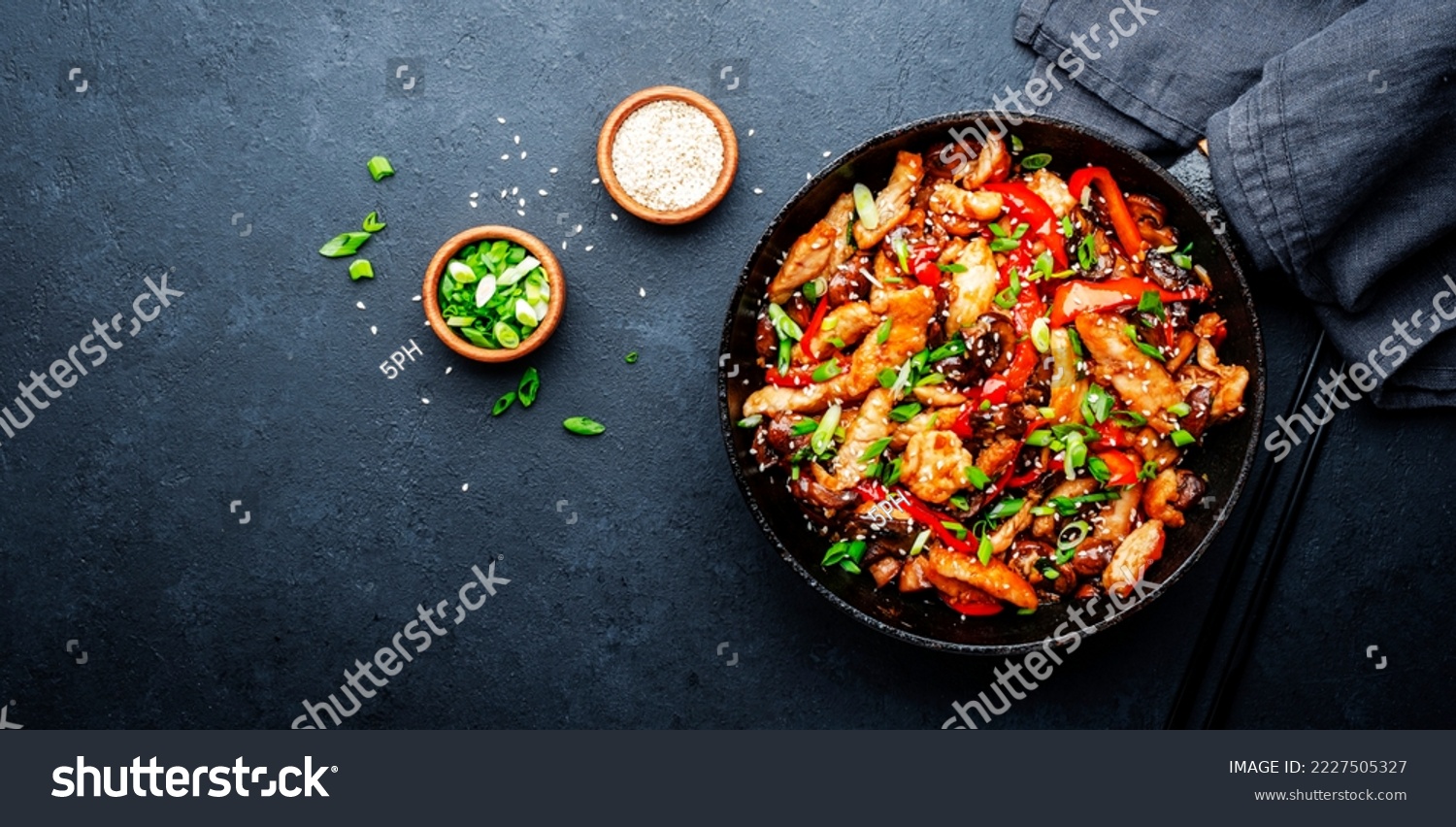 Stir fry with turkey fillet, paprika, mushrooms, green chives and sesame seeds in frying pan. Asian cuisine dish. Black stone kitchen table background, top view #2227505327