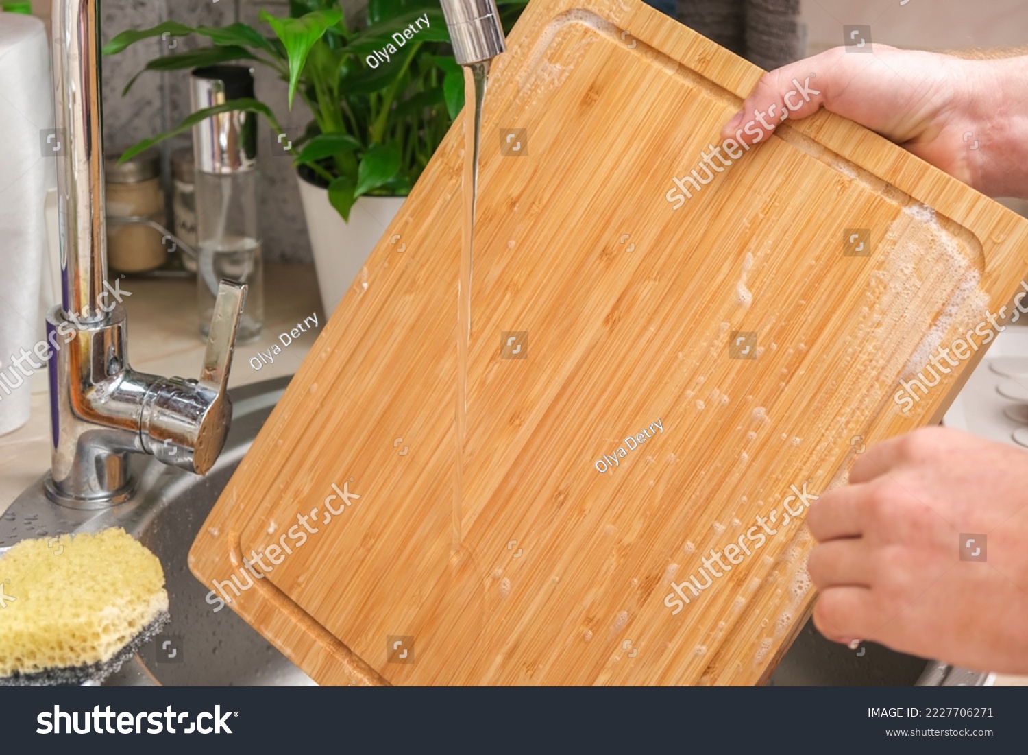 A man washes a wooden bamboo cutting board in the kitchen sink under running water. Gentle hand washing of a wooden cutting board. Cleaning dirty kitchen wood products. #2227706271