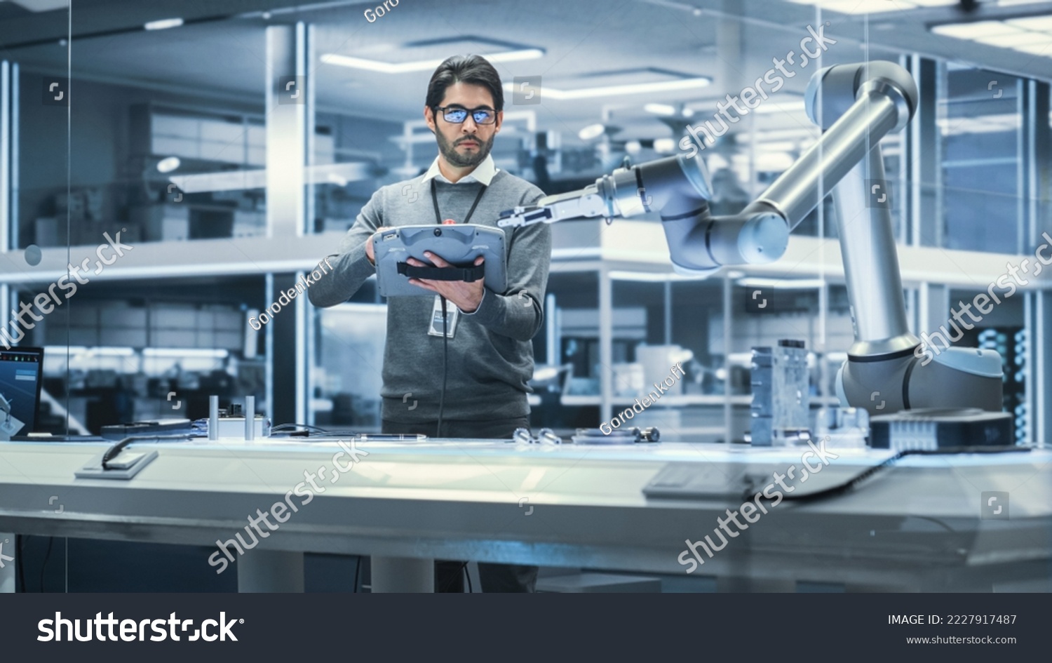 Robotics Engineer Researching and Developing a Technological Robot Arm Project, Standing with Tablet Computer in Scientific Technology Lab. Young Male Working on an AI Robotic Arm. #2227917487