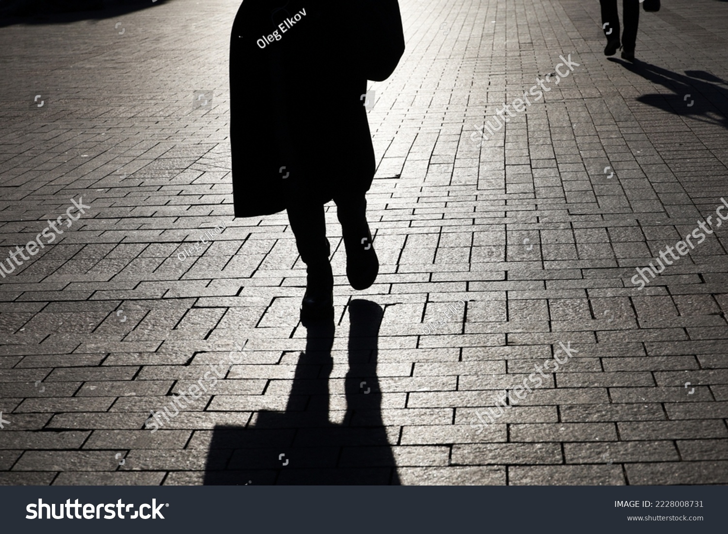 Black silhouette and shadow of lonely woman walking on a street and person on background. Female legs on paved city sidewalk #2228008731