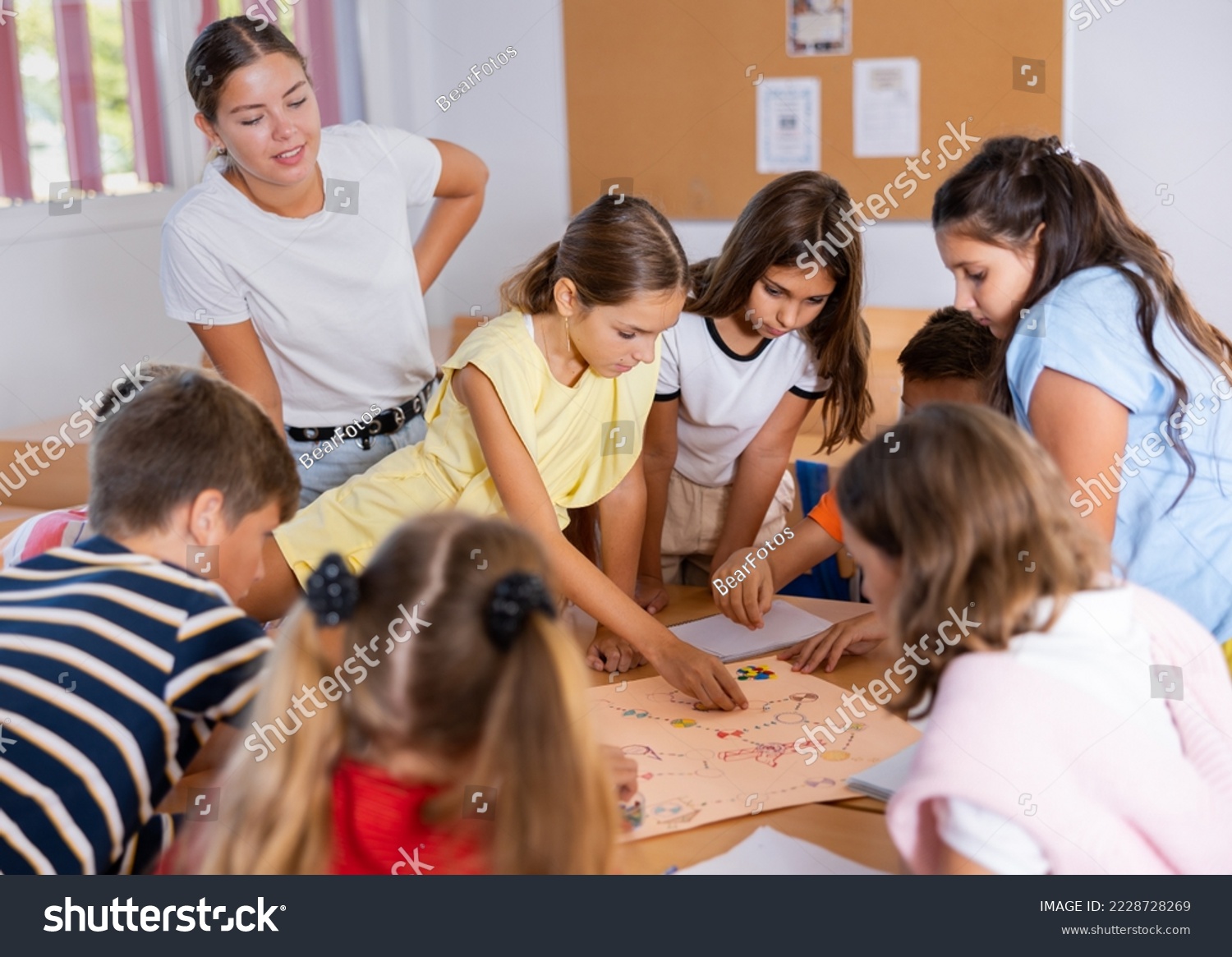 Group of interested school kids with female teacher sitting together around desk in classroom, playing educational tabletop game #2228728269