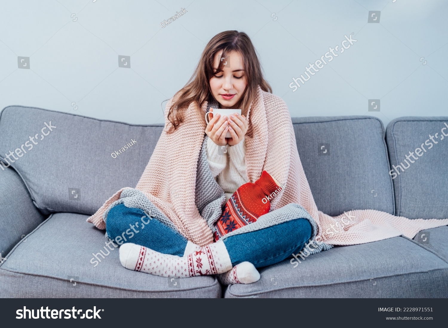 Woman freezes in wintertime. Young girl wearing warm woolen socks and wrapped into two blankets, holding a cup of hot drink and heating pad while sitting on sofa at home. Keep warm. Selective focus. #2228971551