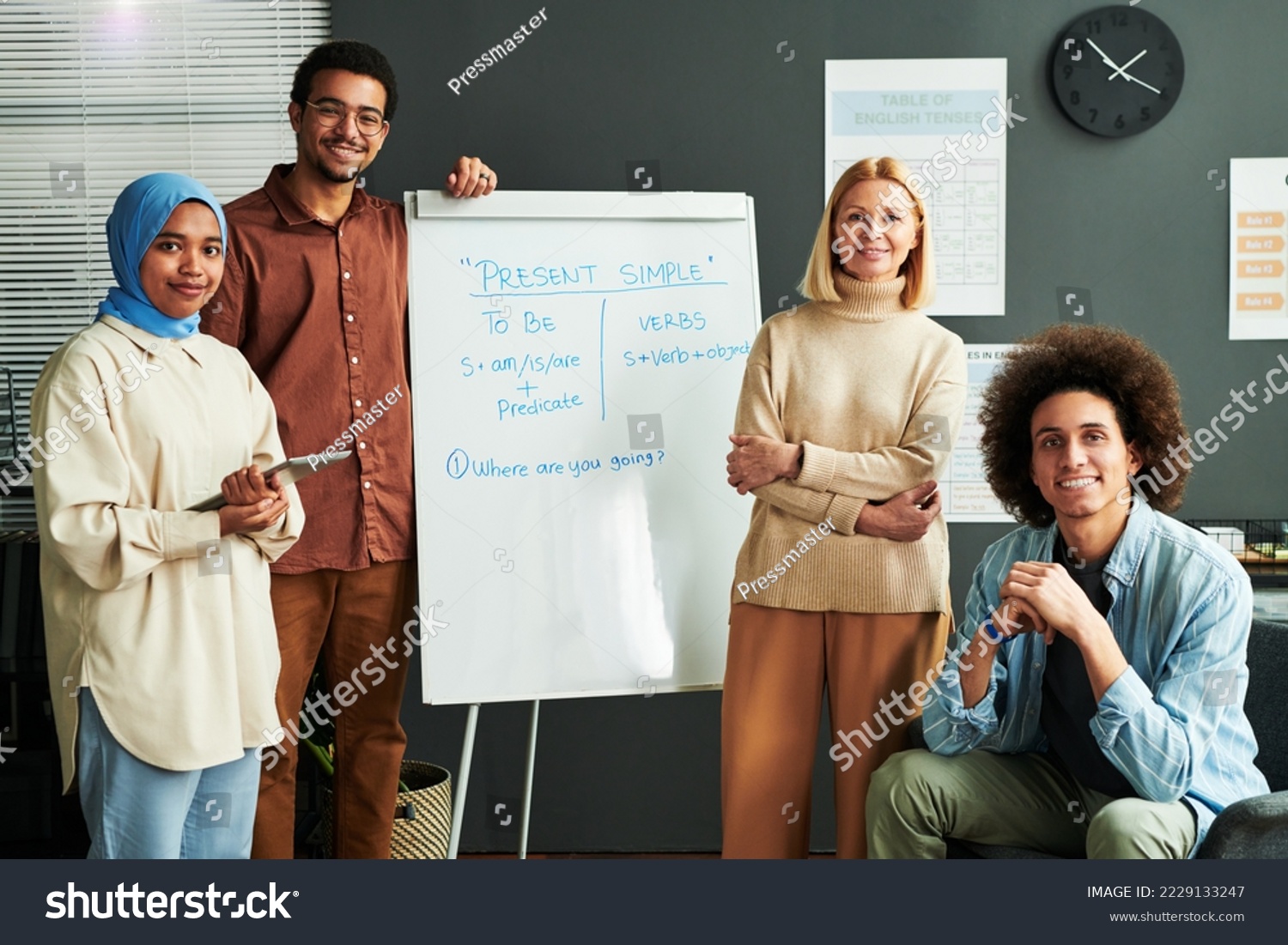 Several happy young intercultural students and mature blond teacher surrounding whiteboard with explanation of Present Simple tense #2229133247