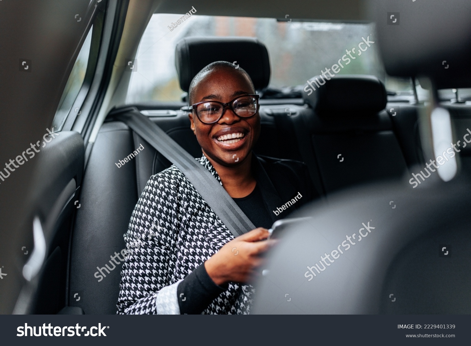 A cheerful African American woman is in the car on the backseat using her smartphone and looking at the camera. #2229401339