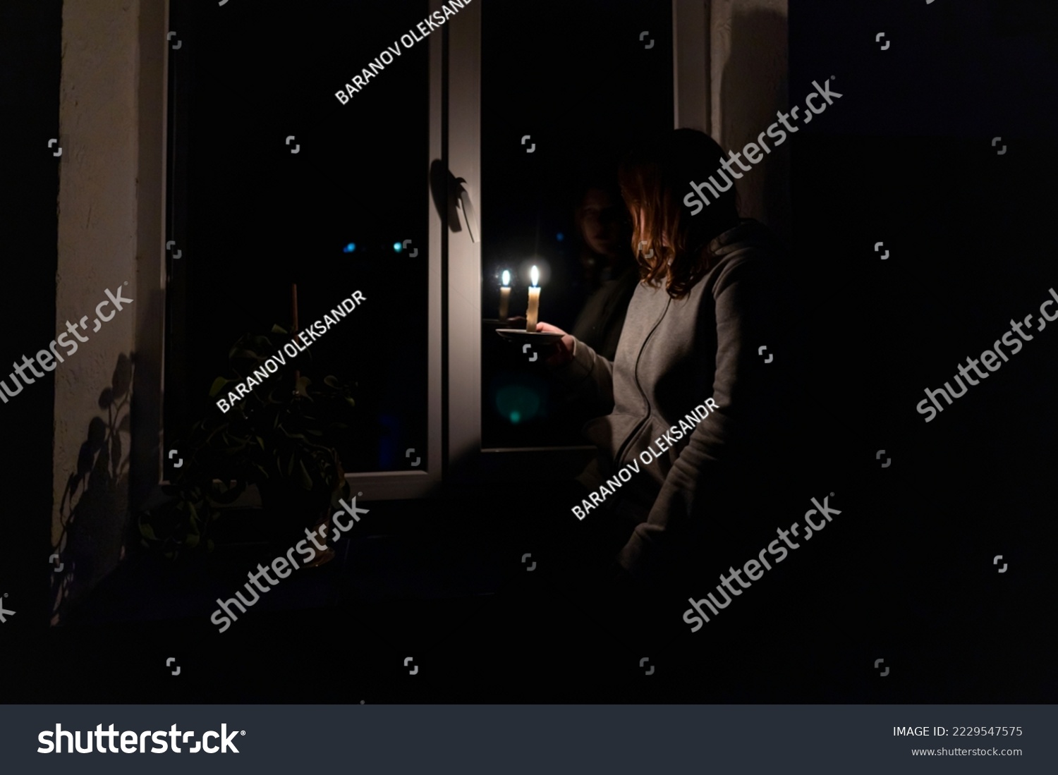 Blackout. Energy crisis. Destruction of infrastructure. Power outage concept. Girl with a burning candle in a dark room sits near the window #2229547575