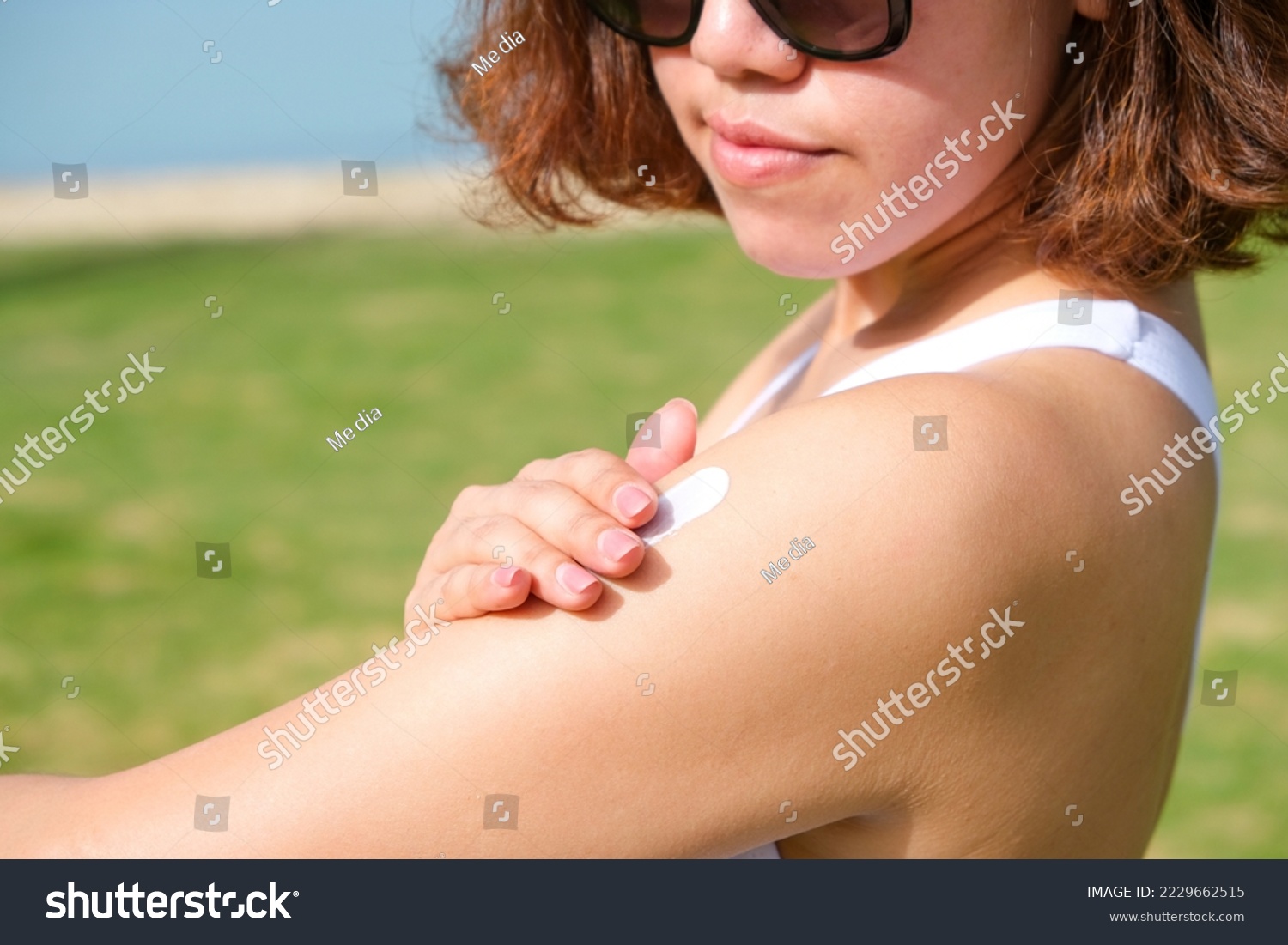A woman is applying sunscreen and skin care to protect her skin from UV rays. She was applying sunscreen to her shoulders and arms. sunny background Health and skin care concept #2229662515