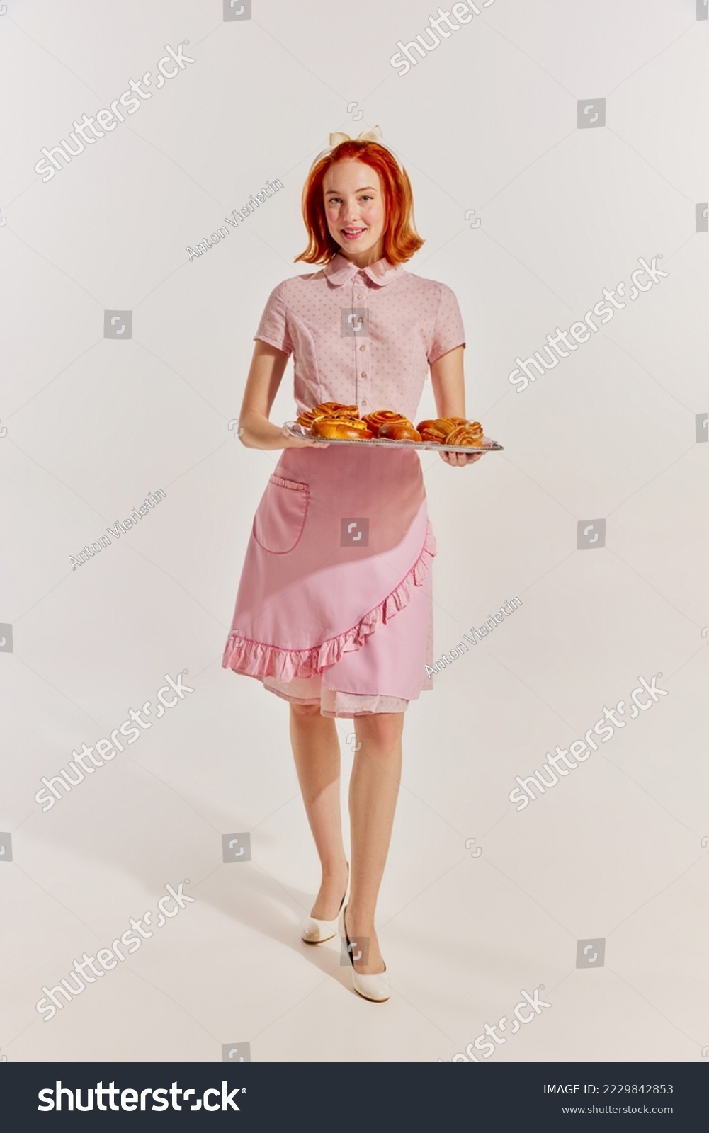 Portrait of young redhead woman in cute pink dress with freshly baked buns isolated over grey background. Concept of beauty, retro style, fashion, elegance, 60s, 70s, family. Copy space for ad #2229842853