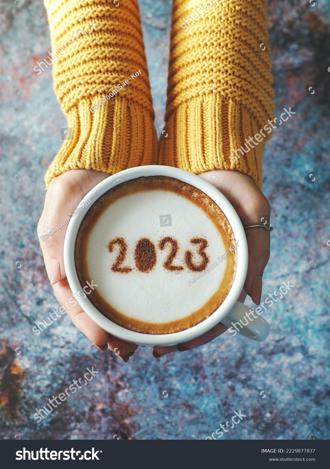 Happy New Year 2023 theme number 2023 on frothy surface of cappuccino served in white coffee mug holding by female hands over rustic blue background. Holidays food art, new year new you. (top view) #2229877837