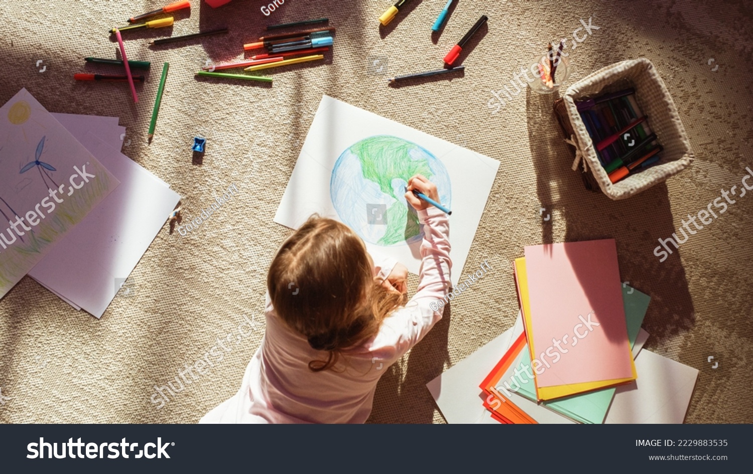 Top View: Little Girl Drawing Our Beautiful Green Planet Earth. Child Having Fun at Home on the Floor, Imagining Our Planet as a Happy Place with Clean, Sustainable Living. Cozy Sunny Day. #2229883535