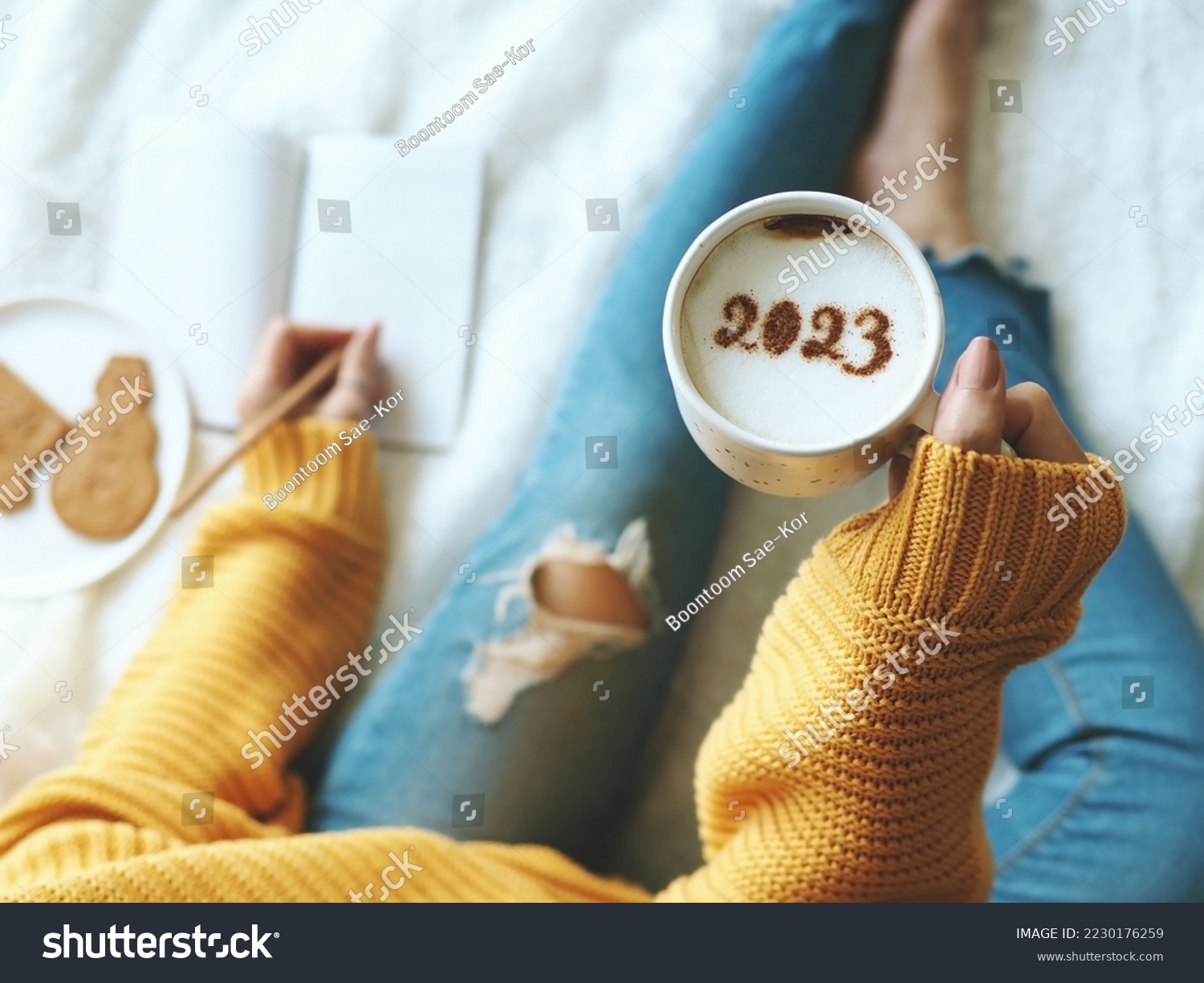 New Year's goal setting, number 2023 on frothy surface of cappuccino in white coffee cup holding by woman in yellow knitted sweater with jeans sitting on bed while writing down her resolutions. #2230176259
