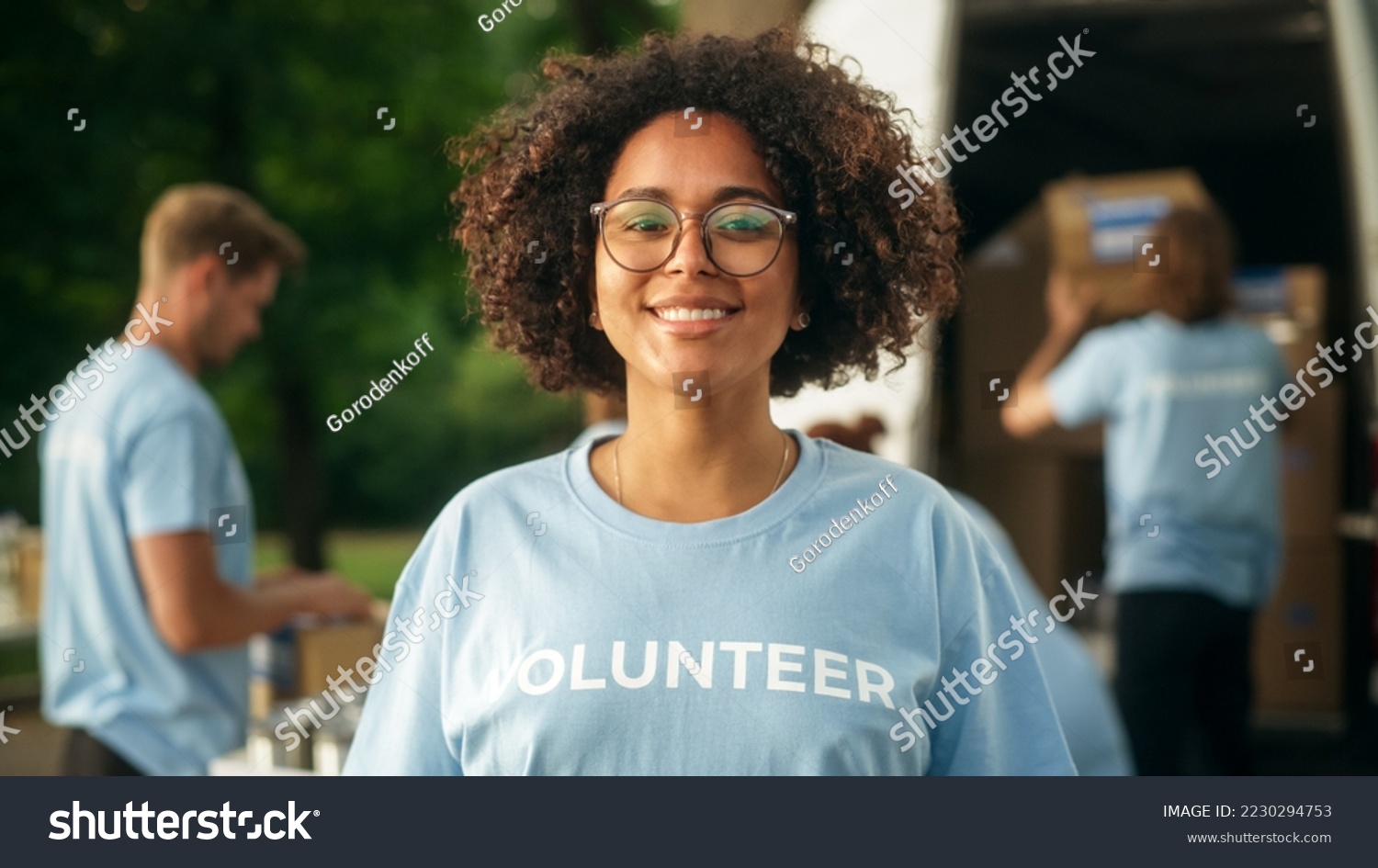 Portrait of a Happy Helpful Black Female Volunteer. Young Adult Multiethnic Latina with Afro Hair, Wearing Glasses, Smiling, Posing for Camera. Humanitarian Aid and Volunteering Concept. #2230294753