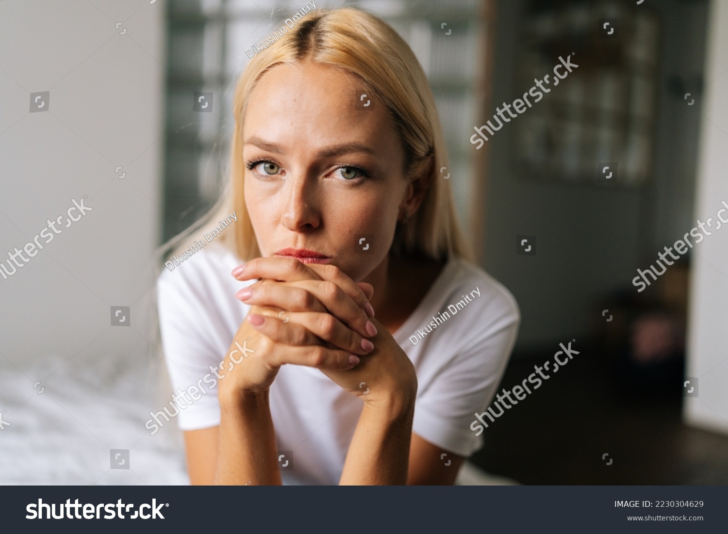 Close up face of thoughtful blonde female sitting alone in living room and serious looking at camera holding hands on chin, thinking over health problems, feeling sadness, boredom, apathy. #2230304629