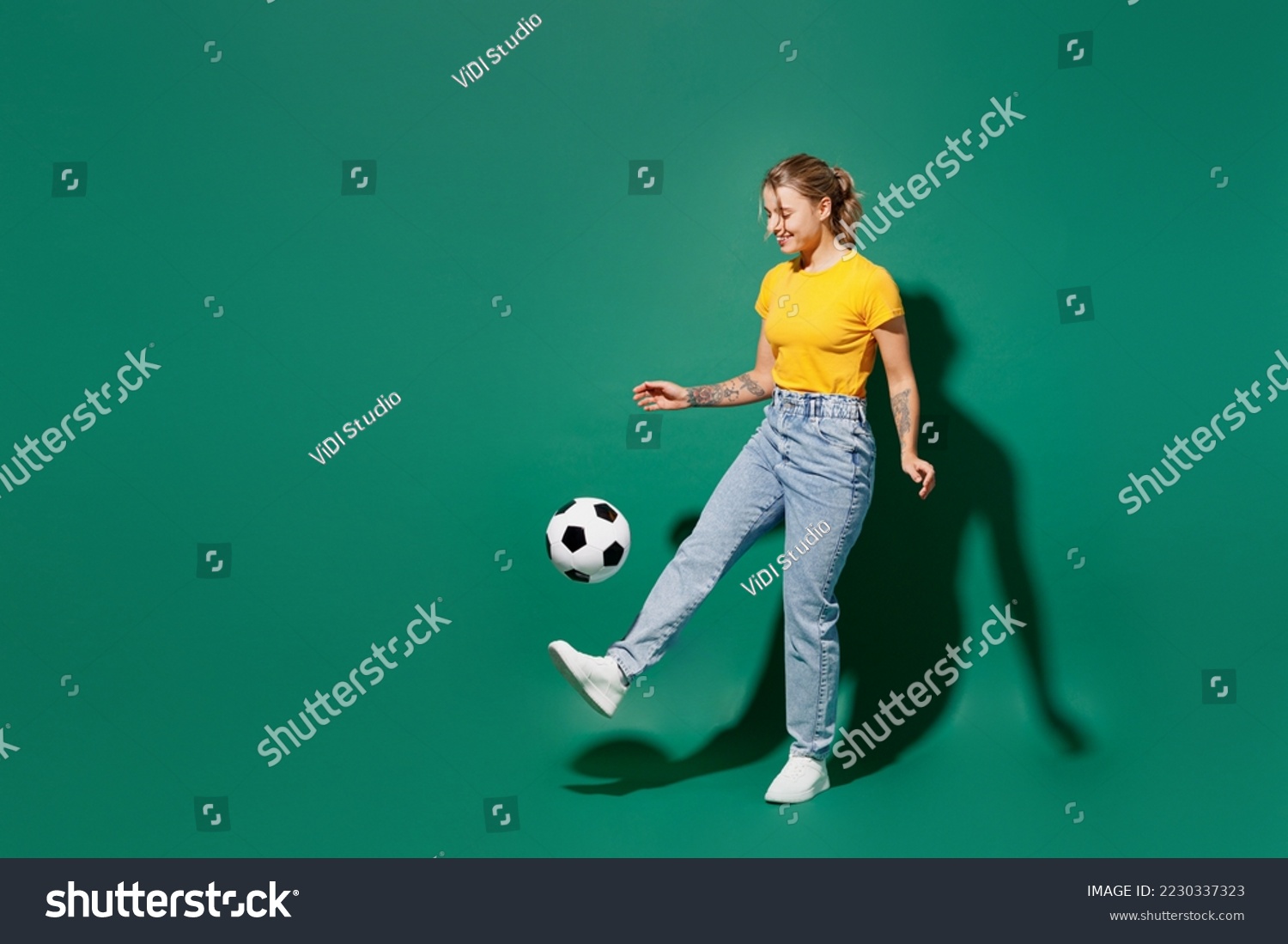 Full body side view fun active young woman fan wear basic yellow t-shirt cheer up support football sport team kick soccer ball on foot leg watch tv live stream isolated on dark green background studio #2230337323