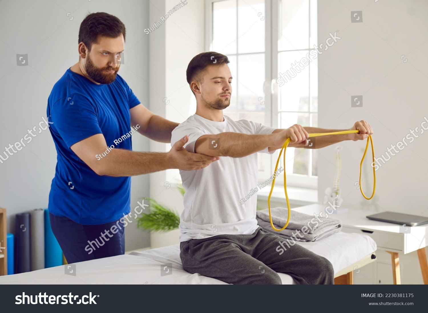 Physiotherapy specialist and his male patient use special equipment during rehabilitation. Young man doing remedial physical exercises with resistance band under supervision of serious physiotherapist #2230381175