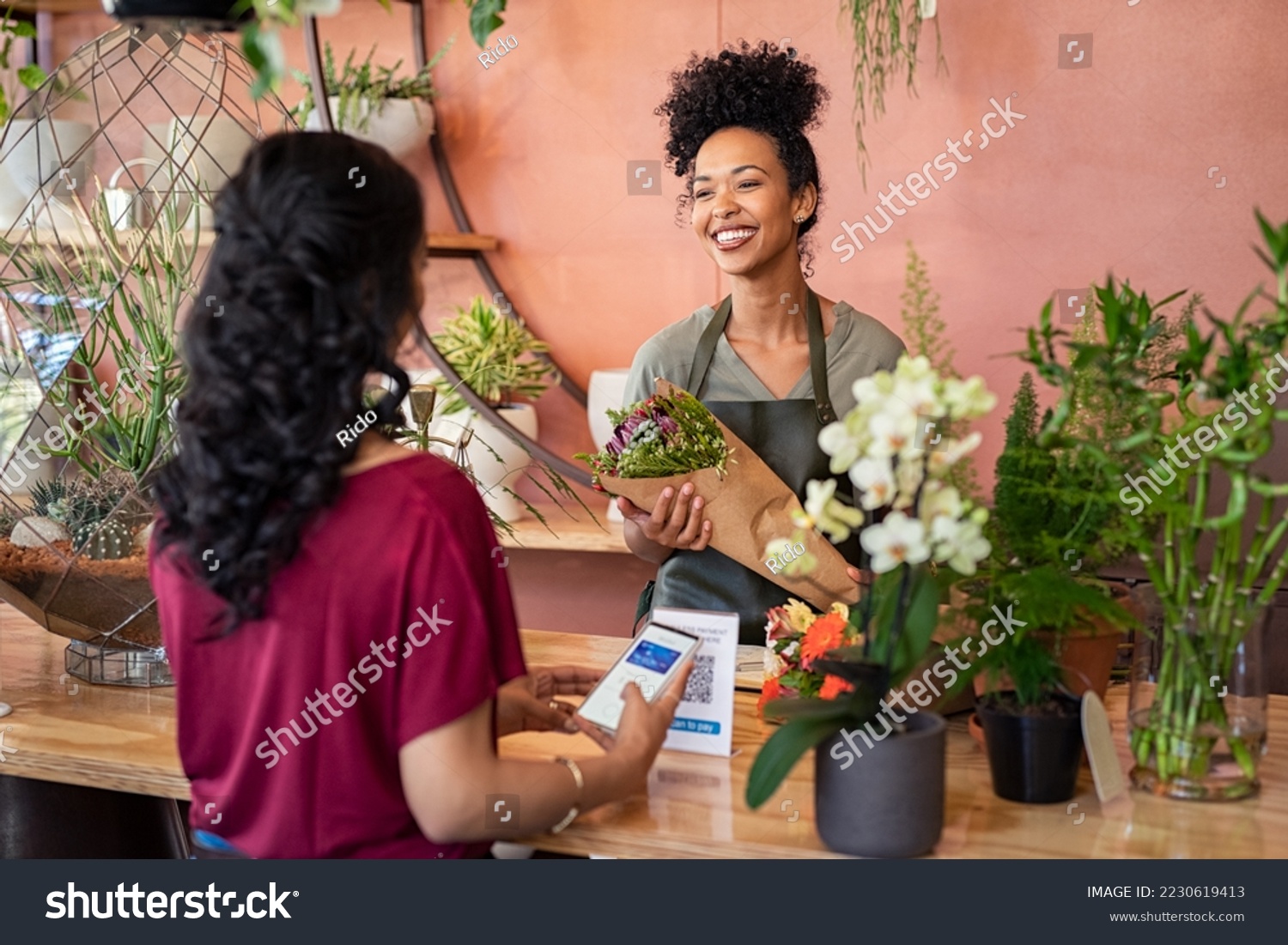 Young african florist holding bouquet of fresh flower while customer making digital payment with smartphone while scanning QR code. Black saleswoman giving a bunch of flower to customer at botany shop #2230619413