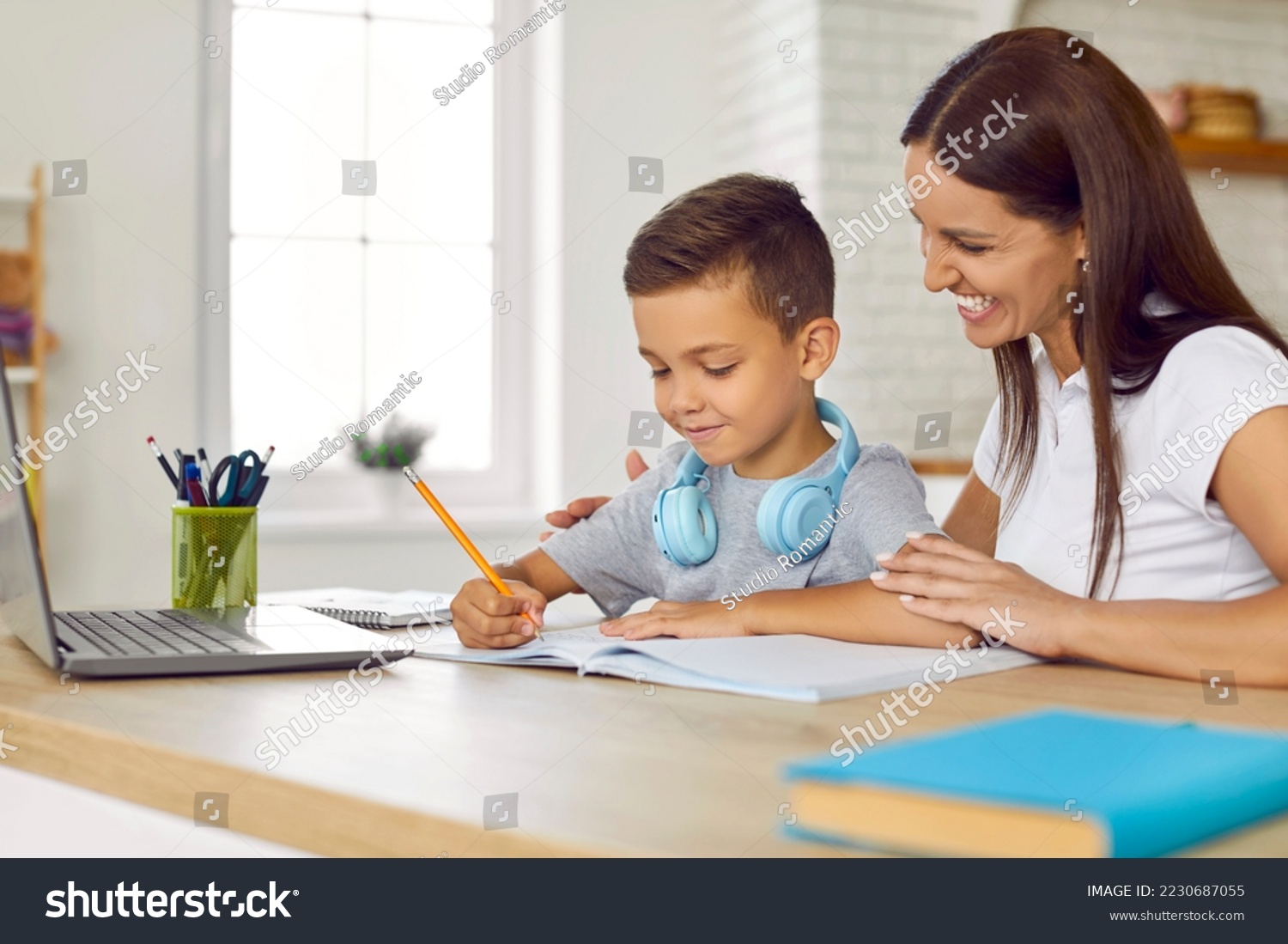 Cheerful family doing school assignment at home. Happy young mother sitting at desk together with her son, helping him with homework, smiling and supporting him. Children, education concept #2230687055