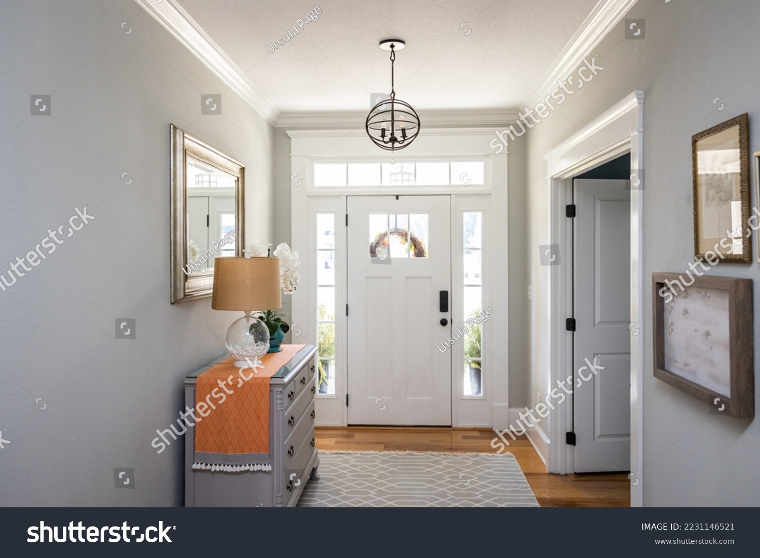 An open large and wide interior front door hallway foyer with transom, hanging light fixture, coastal colors and entry way table and wood floors. #2231146521