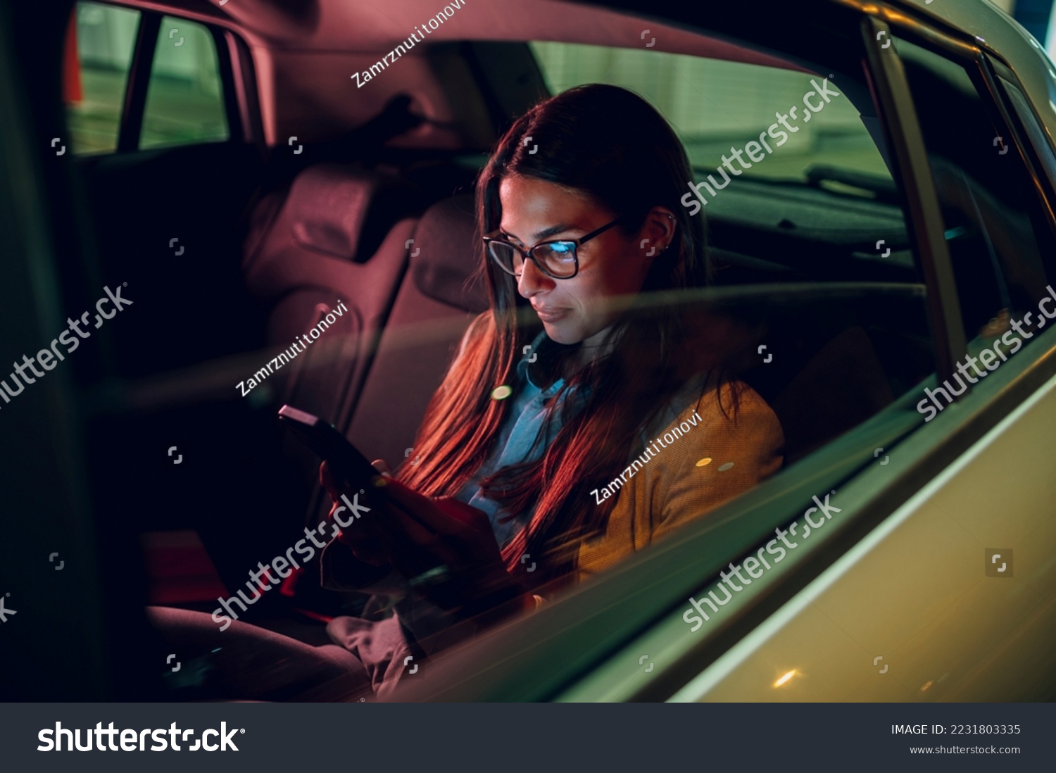 Businesswoman traveling with car on a business trip during night while sitting in a backseat and using a smartphone. Female using mobile phone to send email or messages. View trough car window. #2231803335