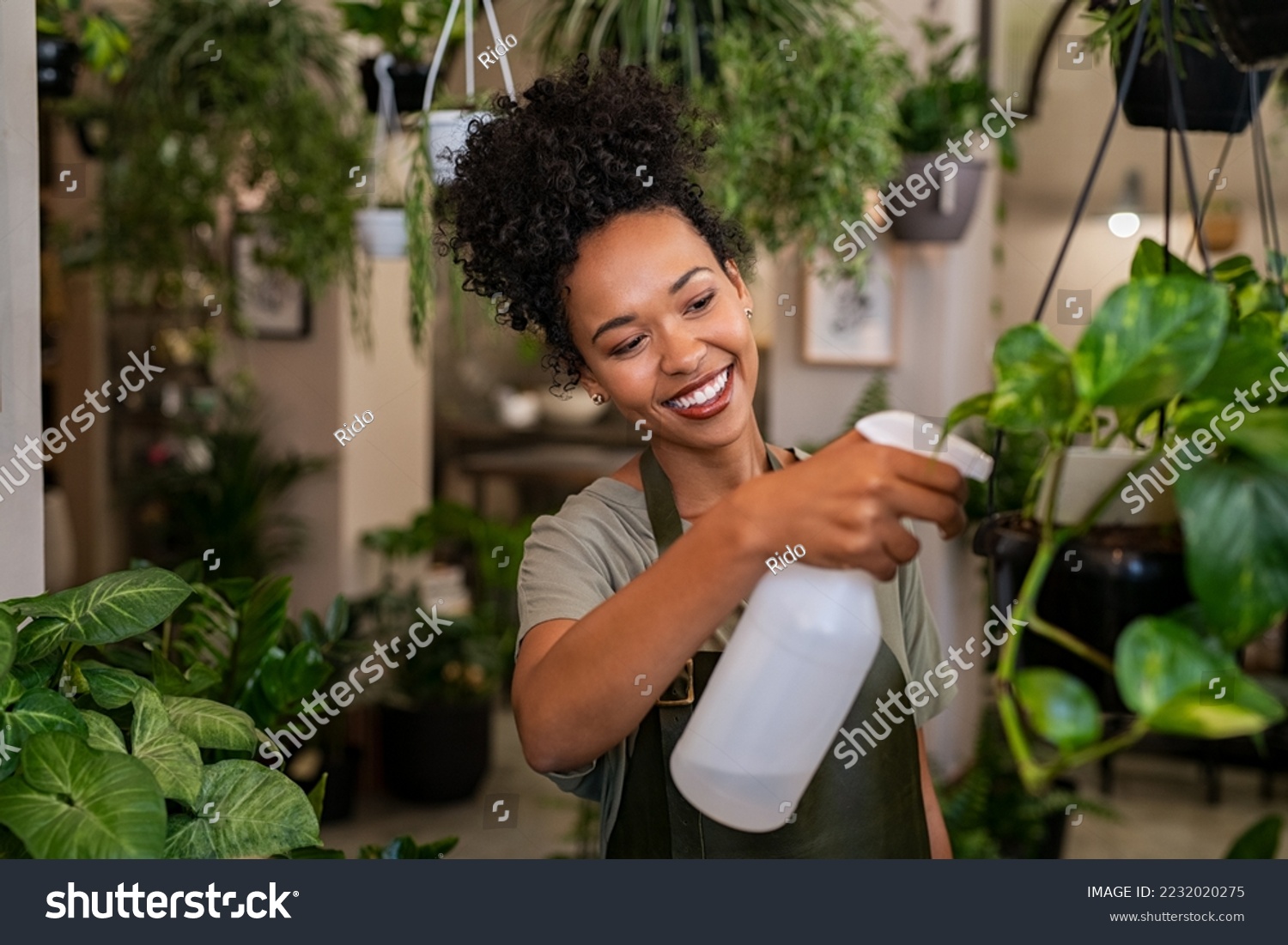 Beautiful black florist taking care of plants while spraying it with water. African american owner working and spraying water plants in store. Happy and smiling florist watering plants in shop. #2232020275