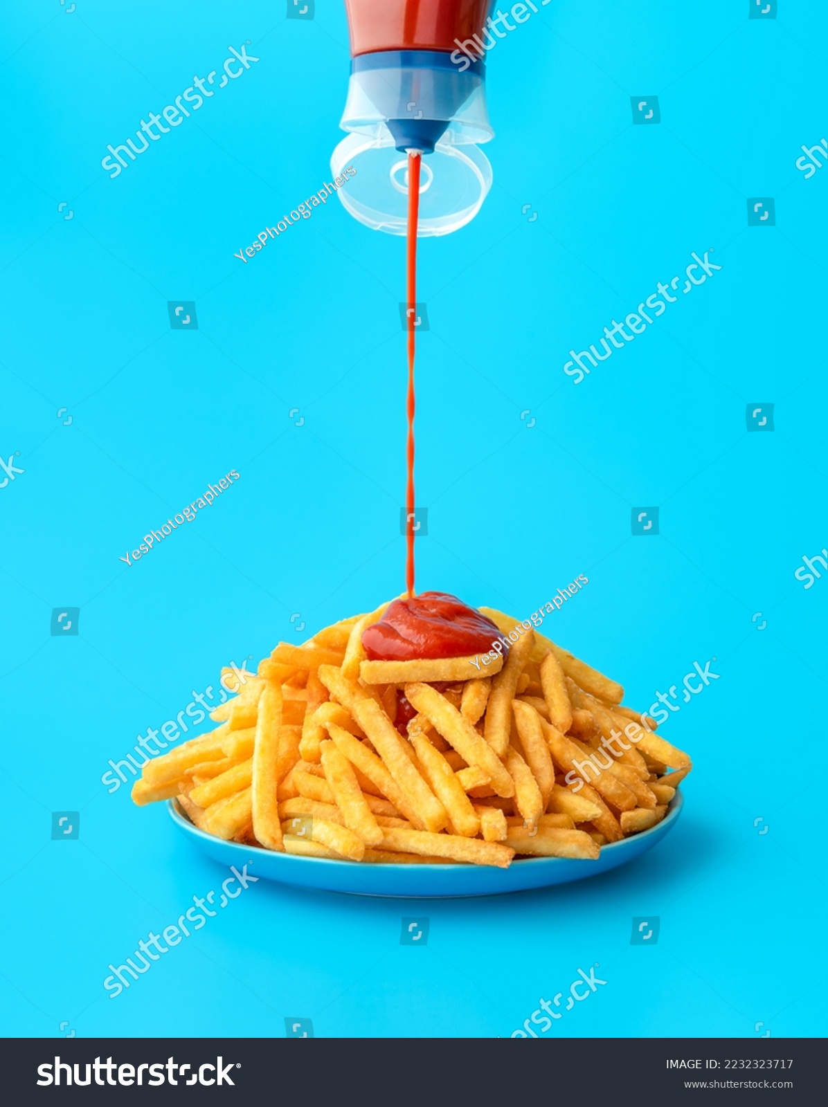 Pouring ketchup from a bottle over french fries, minimalist on a blue background. Delicious meal, french fries with ketchup in bright light on a colorful table. #2232323717